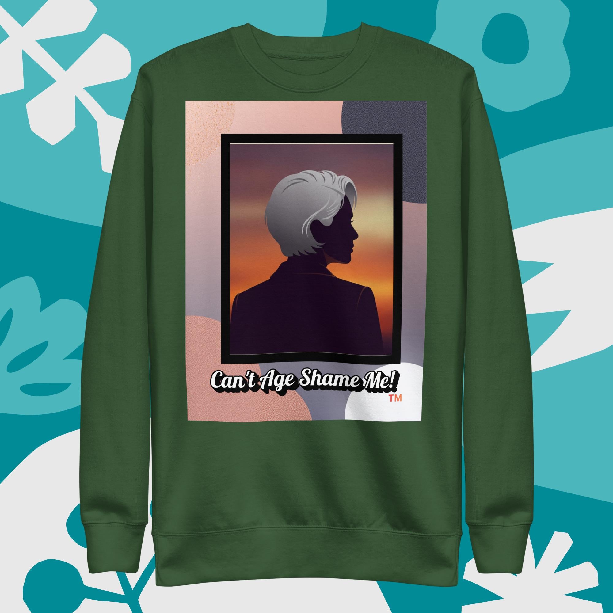 Green sweatshirt with a graphic design featuring a woman with gray hair silhouette and text: Can’t Age Shame Me! surrounded by decorative plants on a light gray background.

