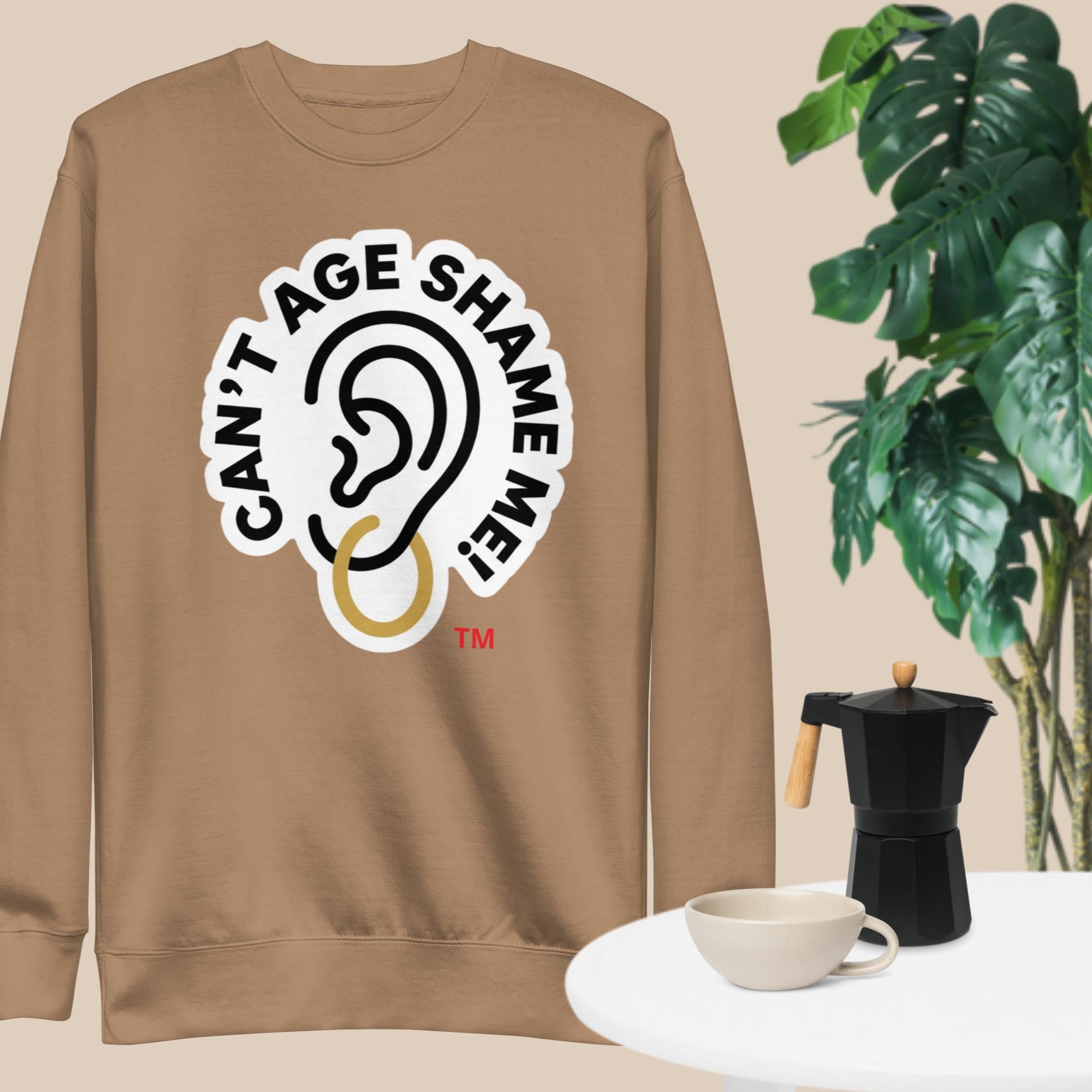 Latte sweatshirt with 'Can't Age Shame Me' graphic on a beige background with a plant and coffee set.