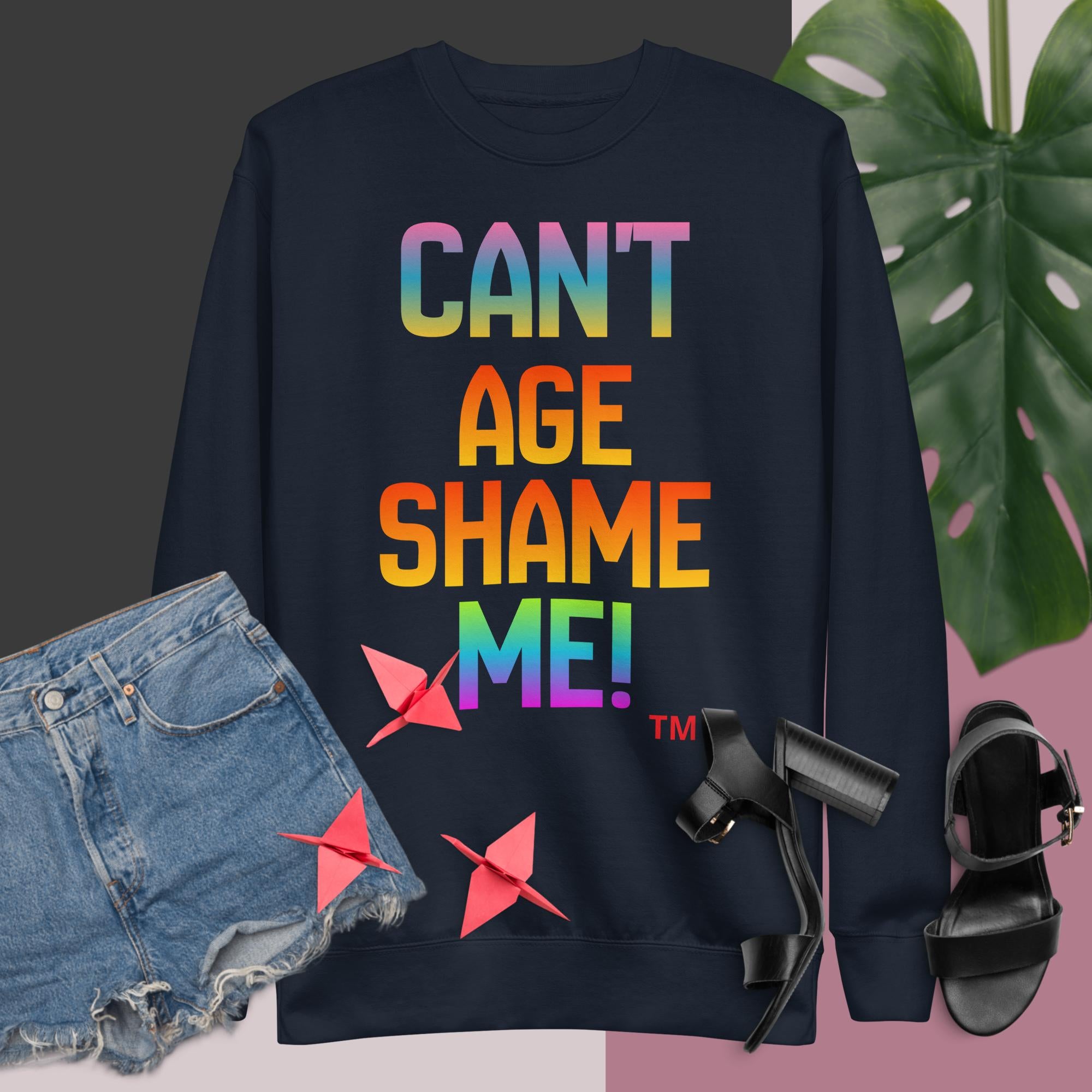 Black sweatshirt with colorful text 'CAN'T AGE SHAME ME!' displayed with denim shorts, sandals, and a plant.