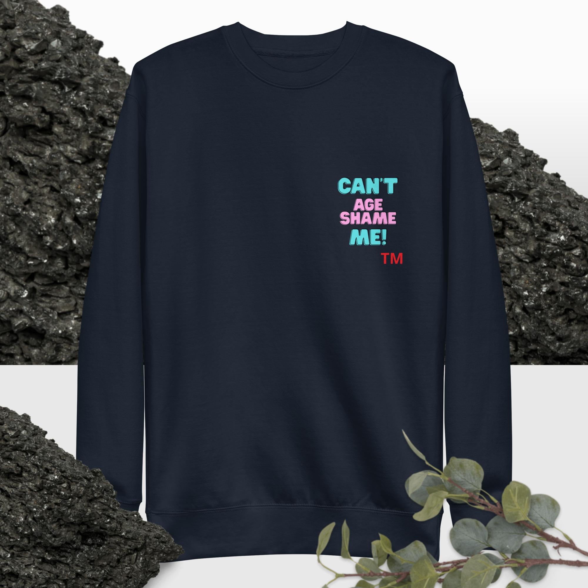 Navy blue sweatshirt with colorful text: 'Can't Age Shame Me'  on a background with black rocks and greenery.