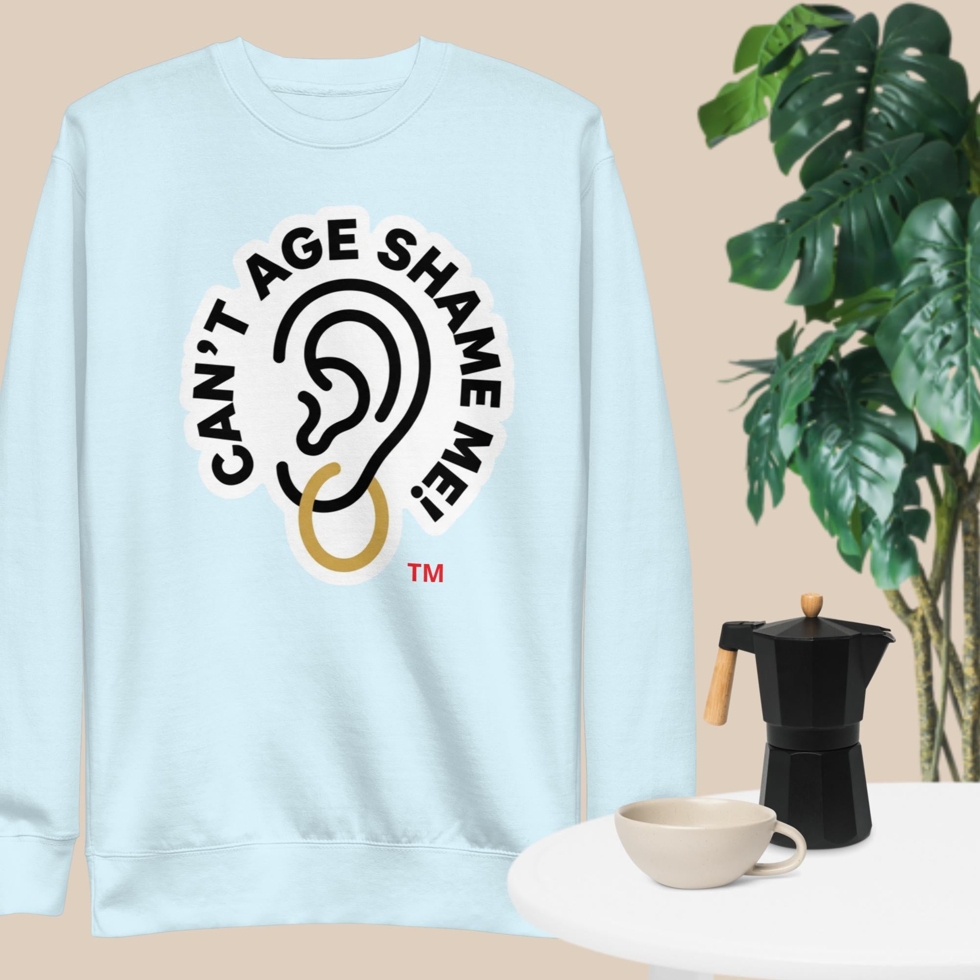 Light blue sweatshirt with 'Can't Age Shame Me!' graphic on a beige background with a plant and coffee maker.