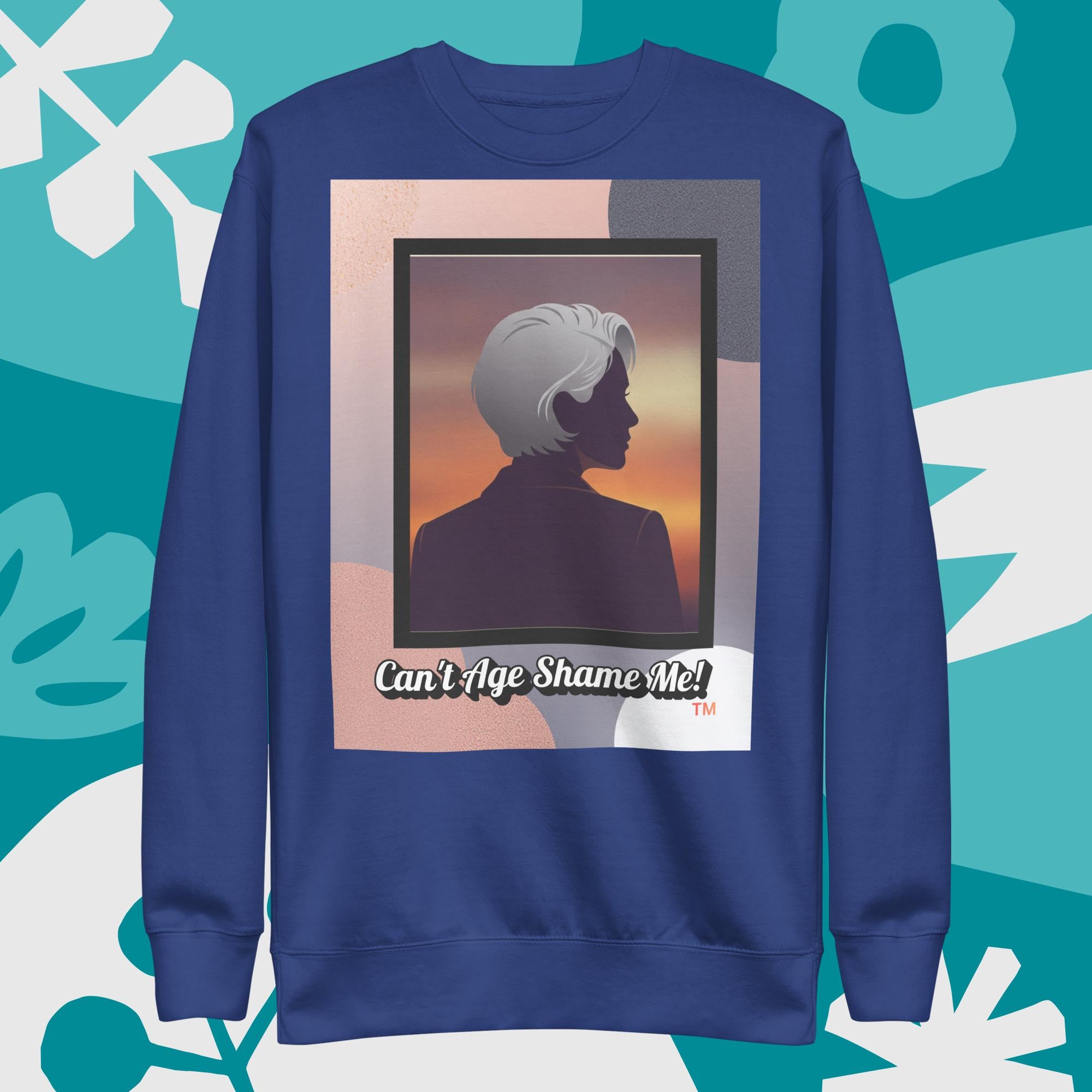 Purplish blue sweatshirt with a graphic design featuring a woman with gray hair silhouette and text: Can’t Age Shame Me! surrounded by decorative plants on a light gray background.

