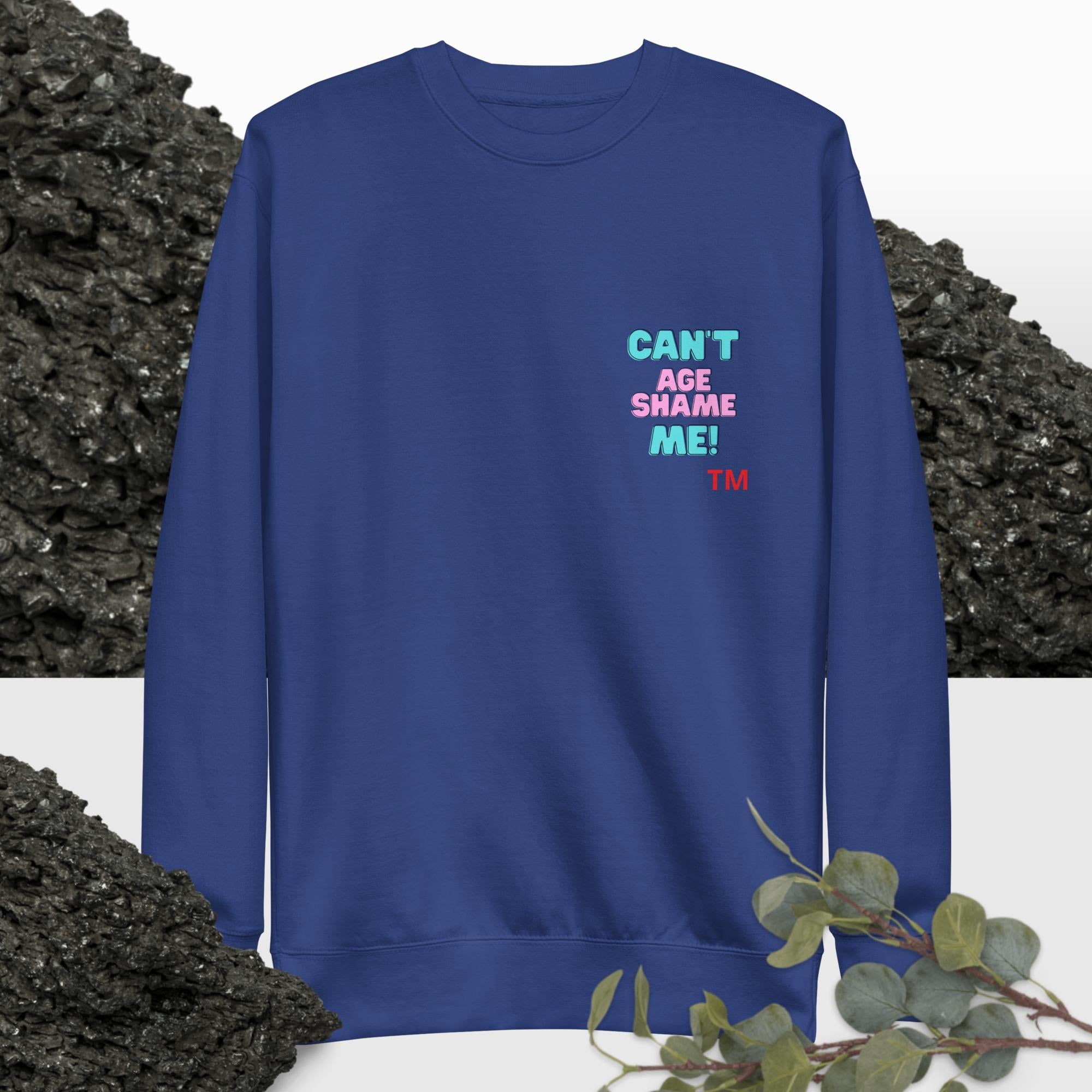 Royal Blue sweatshirt with colorful text: 'Can't Age Shame Me'  on a background with black rocks and greenery.