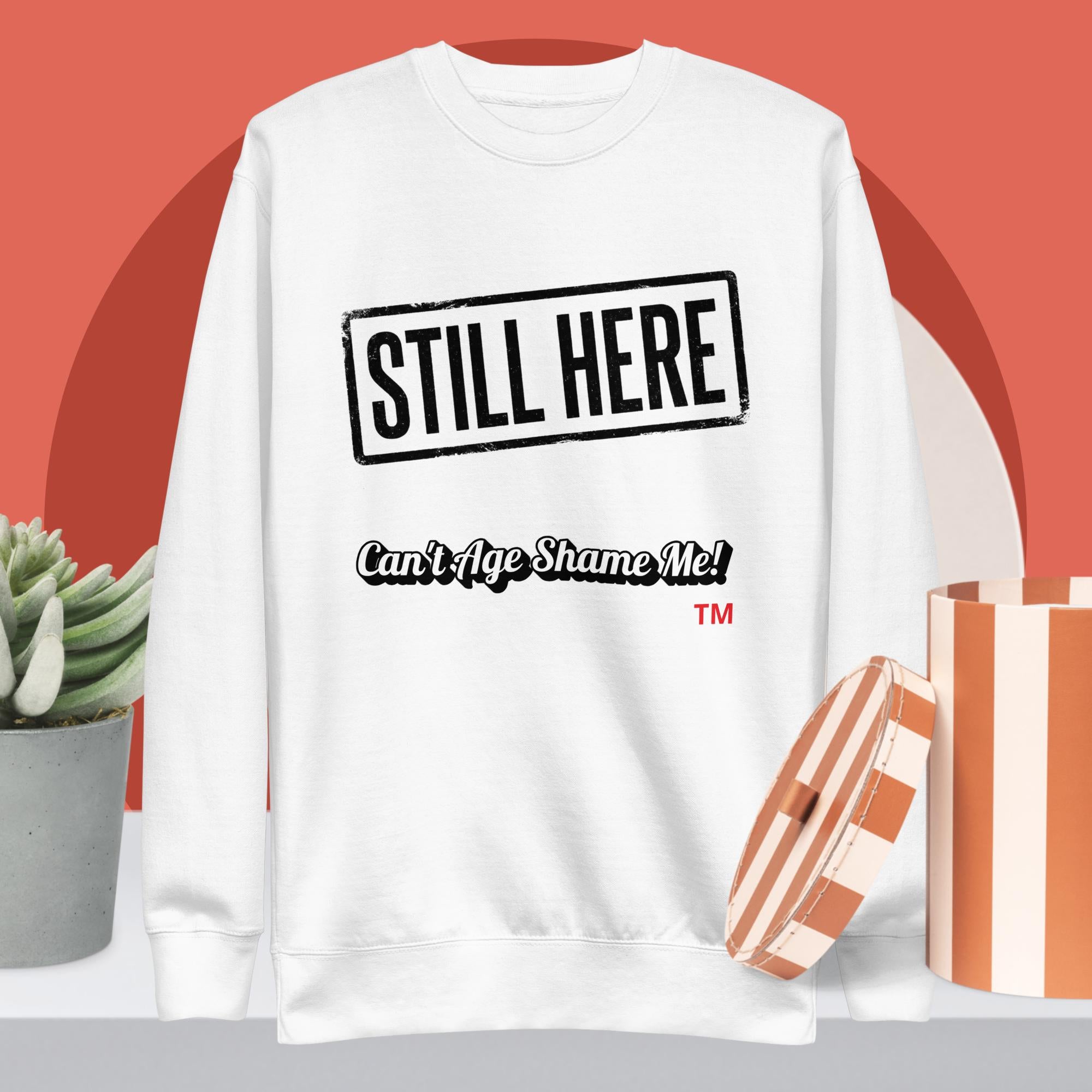 White sweatshirt with 'STILL HERE Can't Age Shame Me!' text on a red background