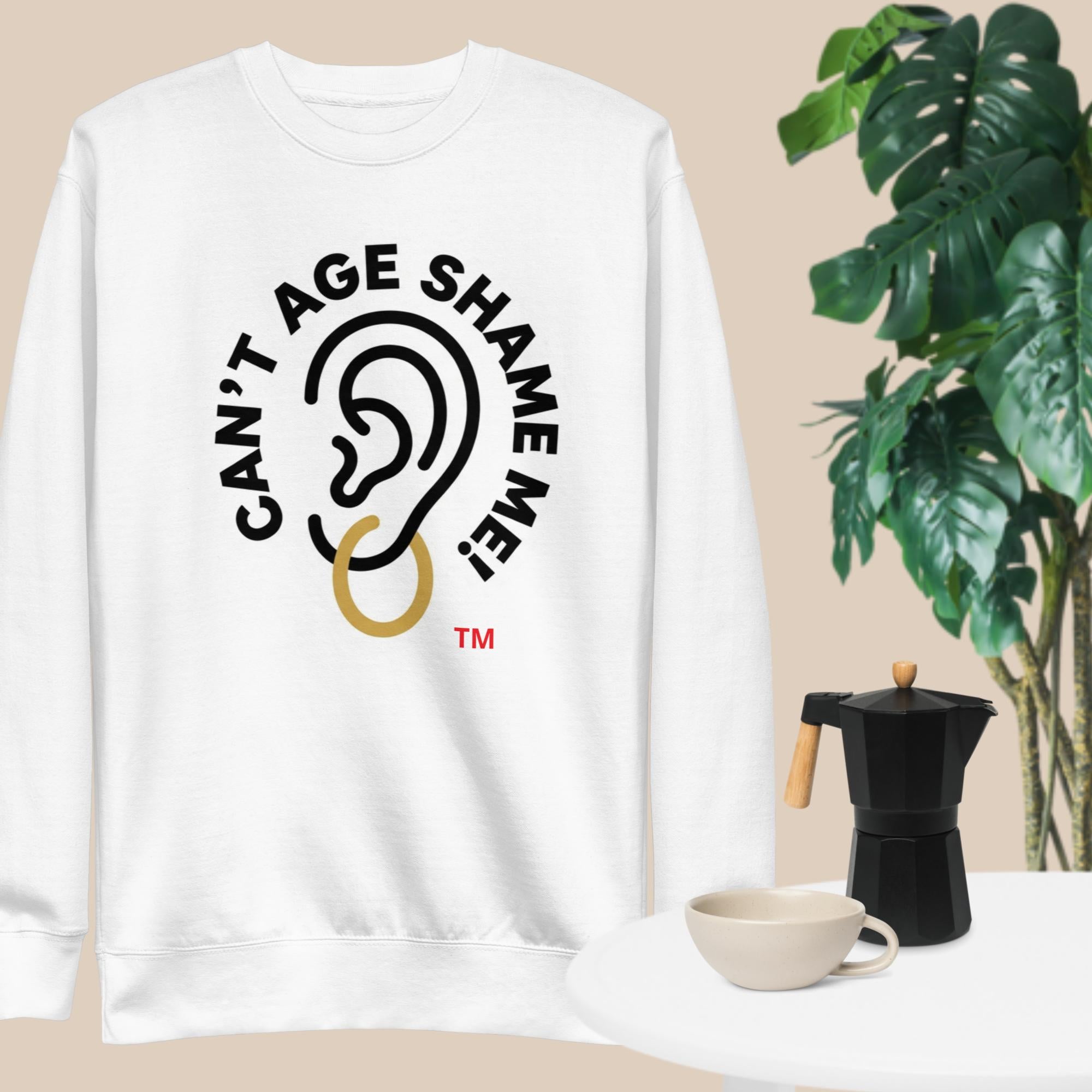 White sweatshirt with 'Can't Age Shame Me' graphic on a beige background with a plant and coffee maker.