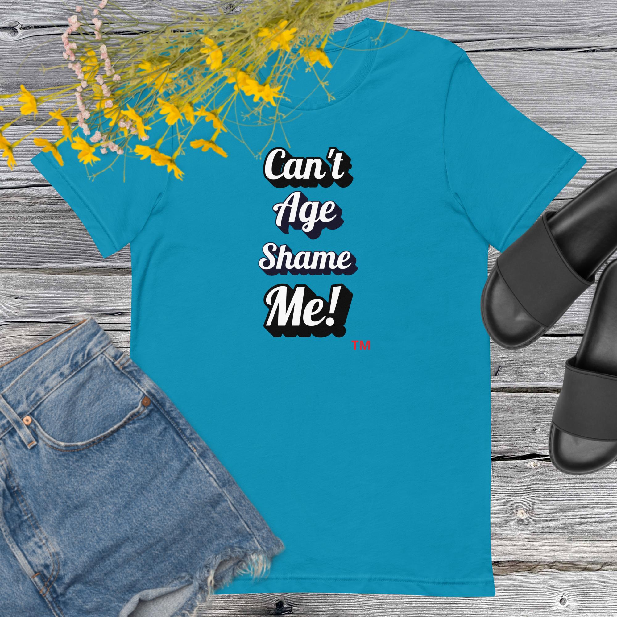 Blue t-shirt with 'Can't Age Shame Me!' text on a wooden surface with denim shorts and sandals.