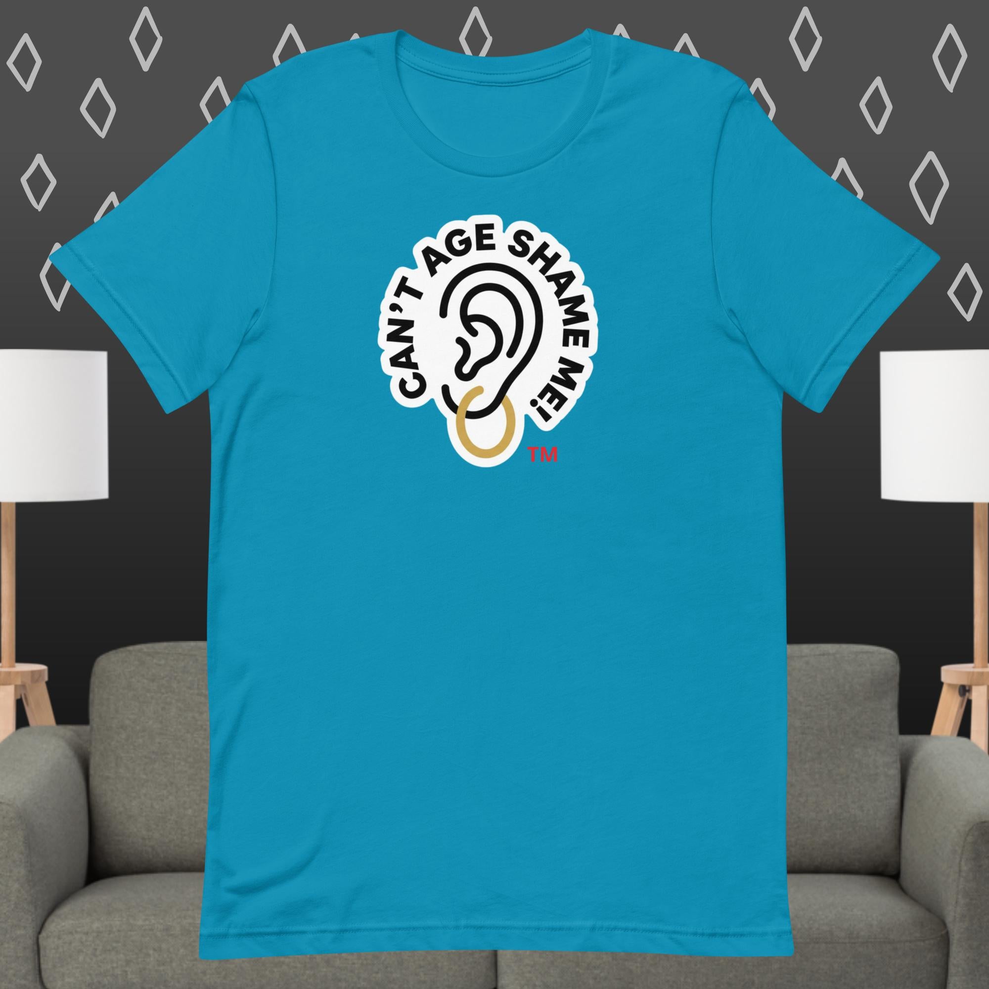 Turquoise t-shirt with 'Can't Age Shame Me' graphic on a gray couch and lamp background