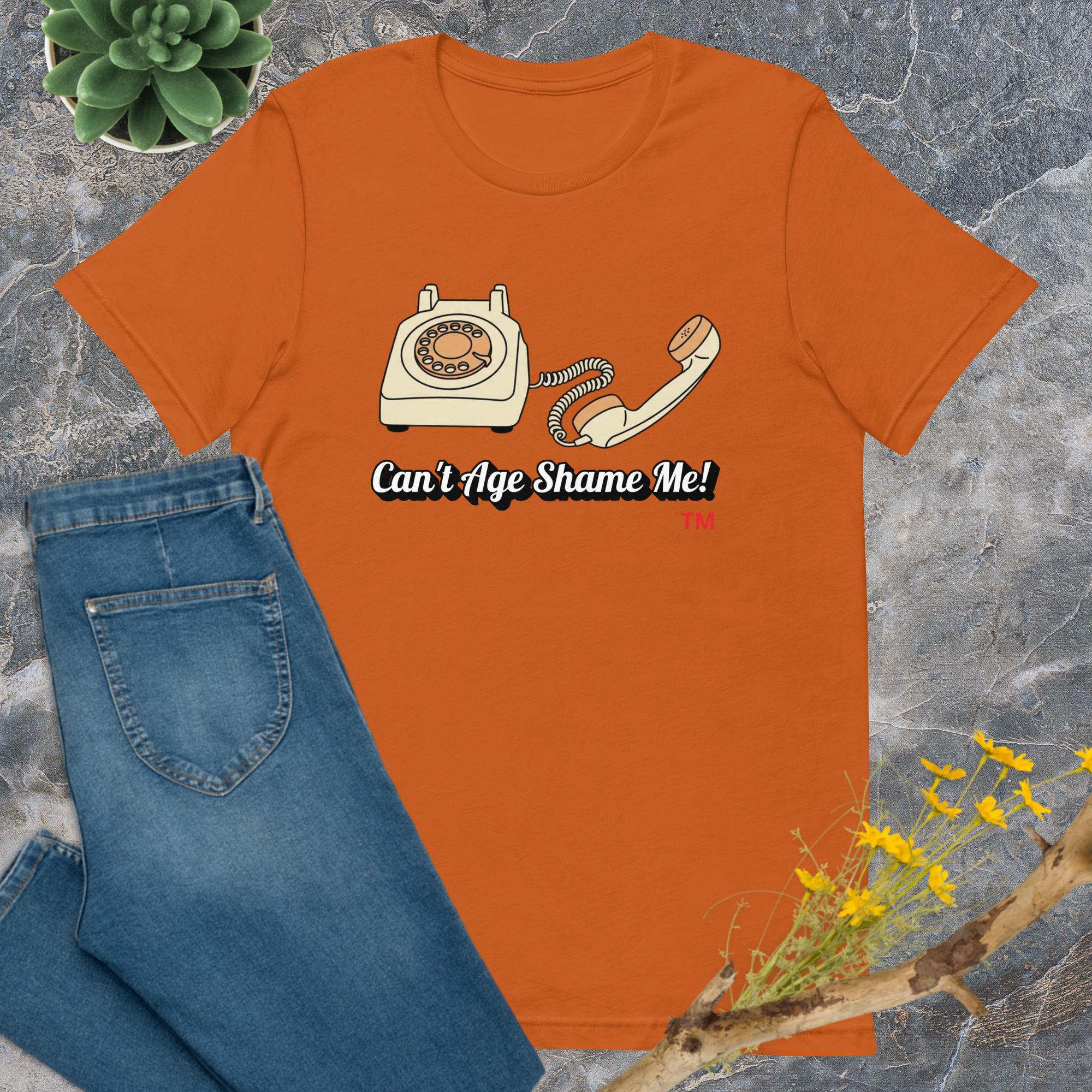 autumn  t-shirt with a phone graphic and text 'Can't Age Shame Me!' on a gray surface with jeans and plants.