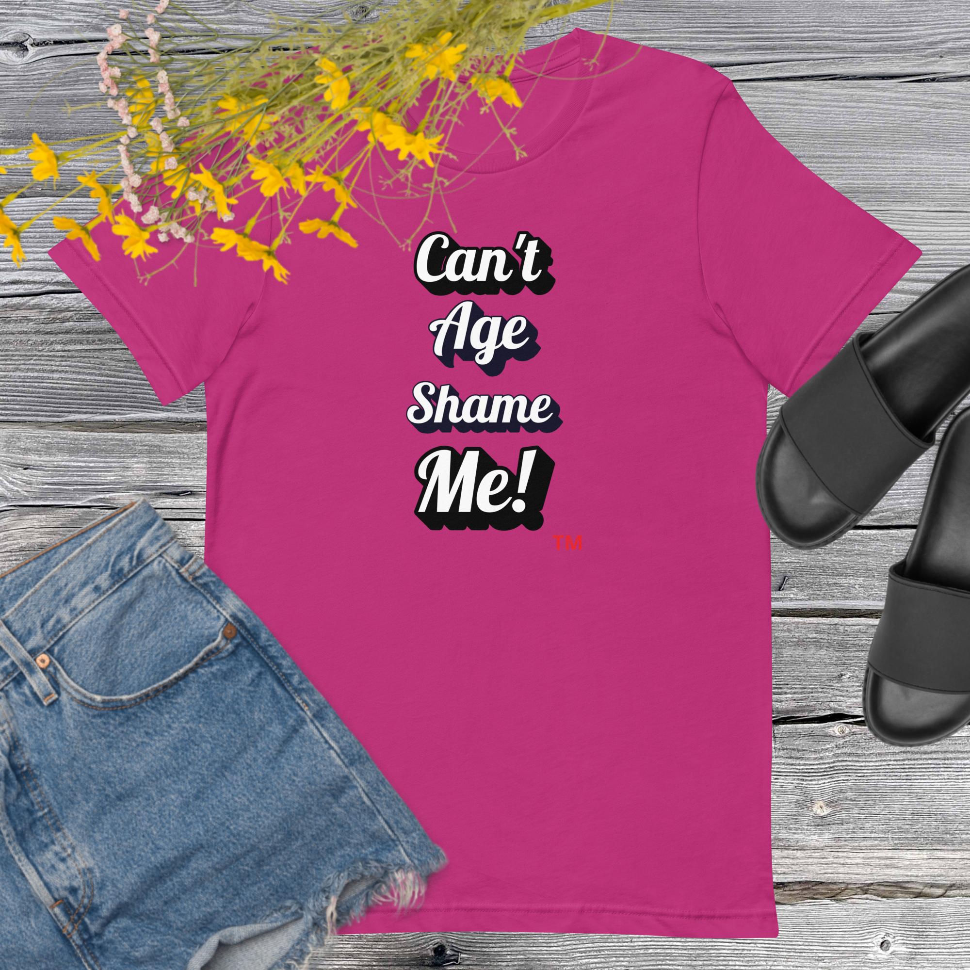 Pink t-shirt with 'Can't Age Shame Me!' text, paired with denim shorts and black sandals on a wooden surface.