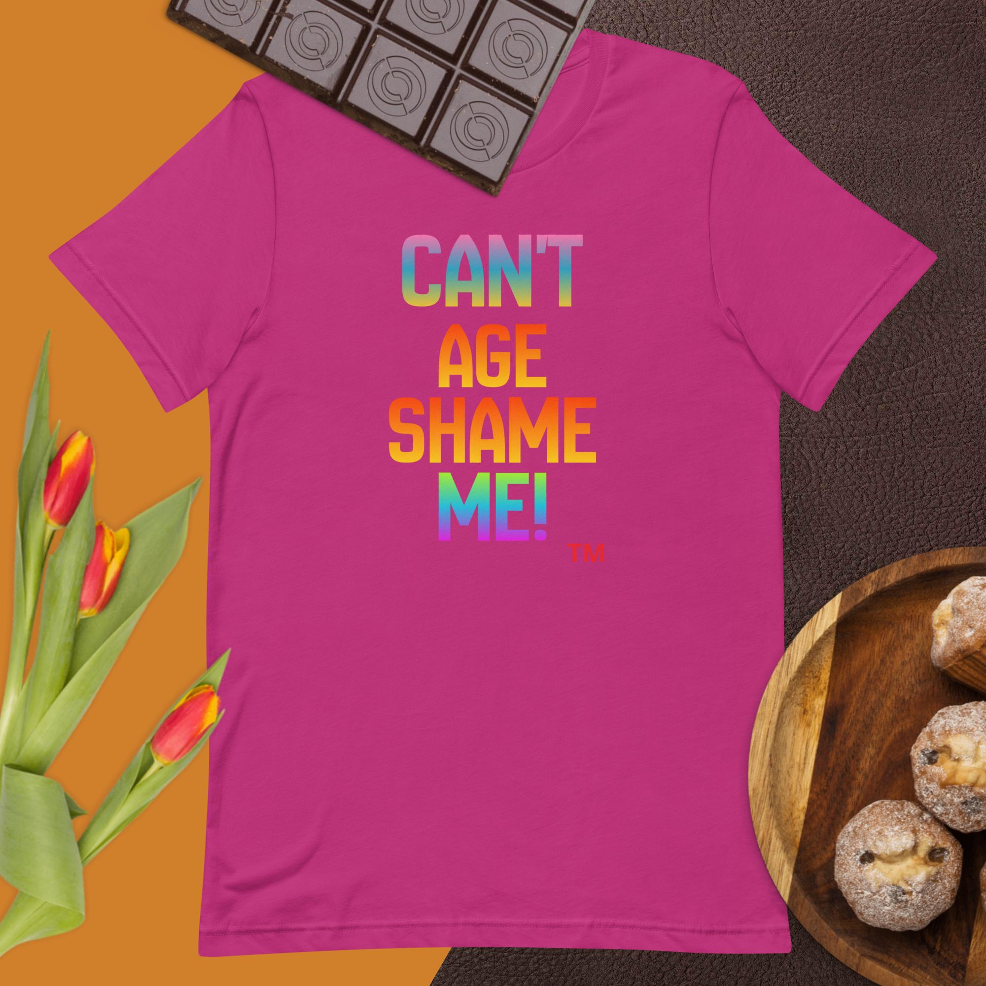 Pink t-shirt with colorful text 'CAN'T AGE SHAME ME!' on a brown surface with tulips and cookies.