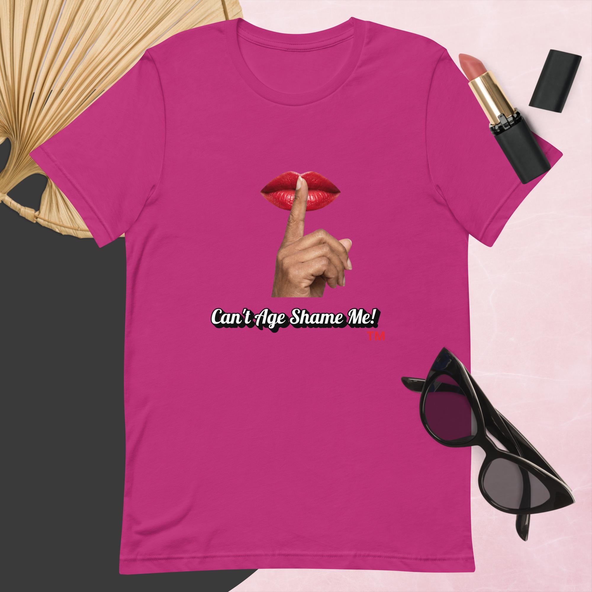 Berry  t-shirt with graphic design and text, Can’t Age Shame Me!,   surrounded by sunglasses, lipstick, and a fan on a pink background.

 
