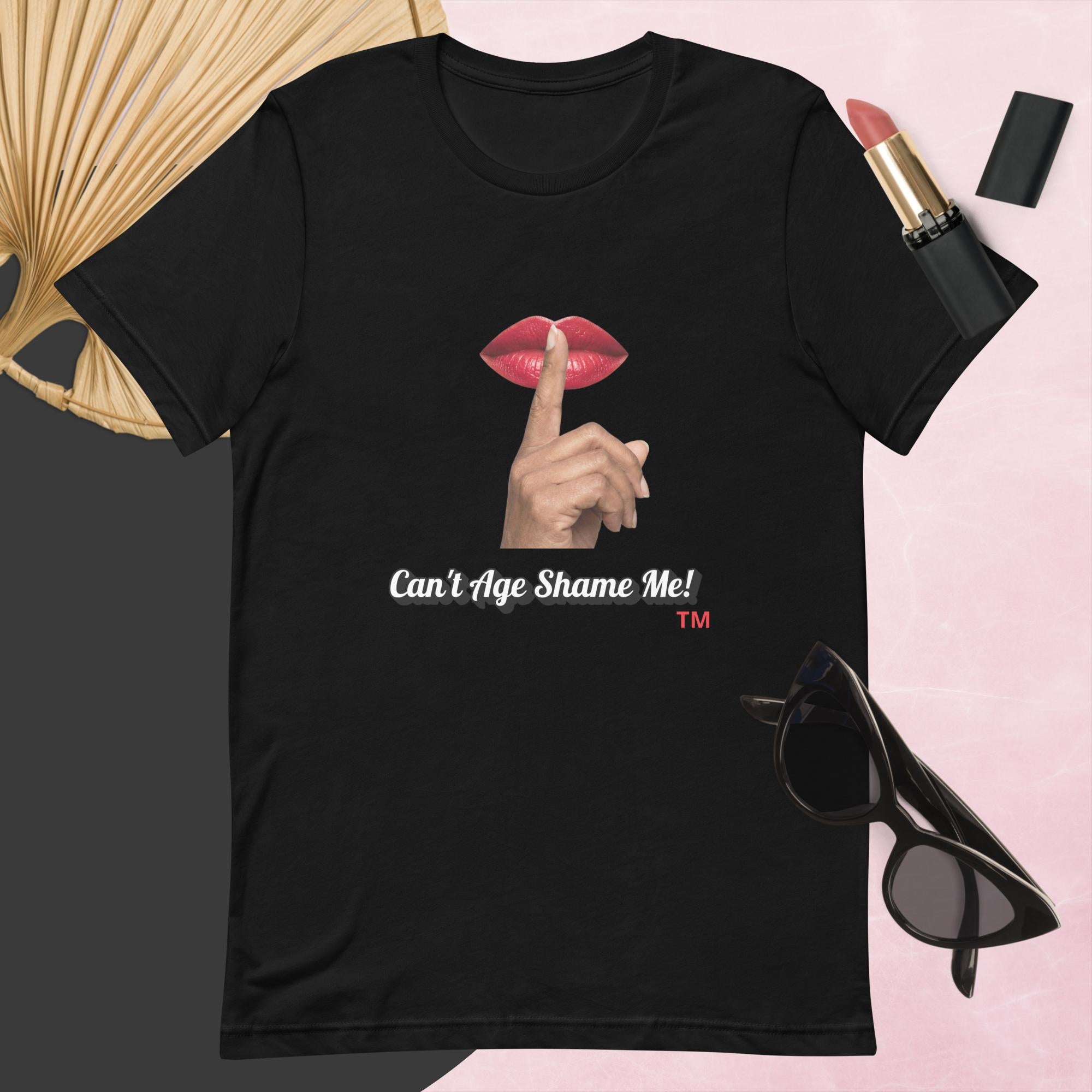 Black  t-shirt with graphic design and text, Can’t Age Shame Me!,   surrounded by sunglasses, lipstick, and a fan on a pink background.

 
