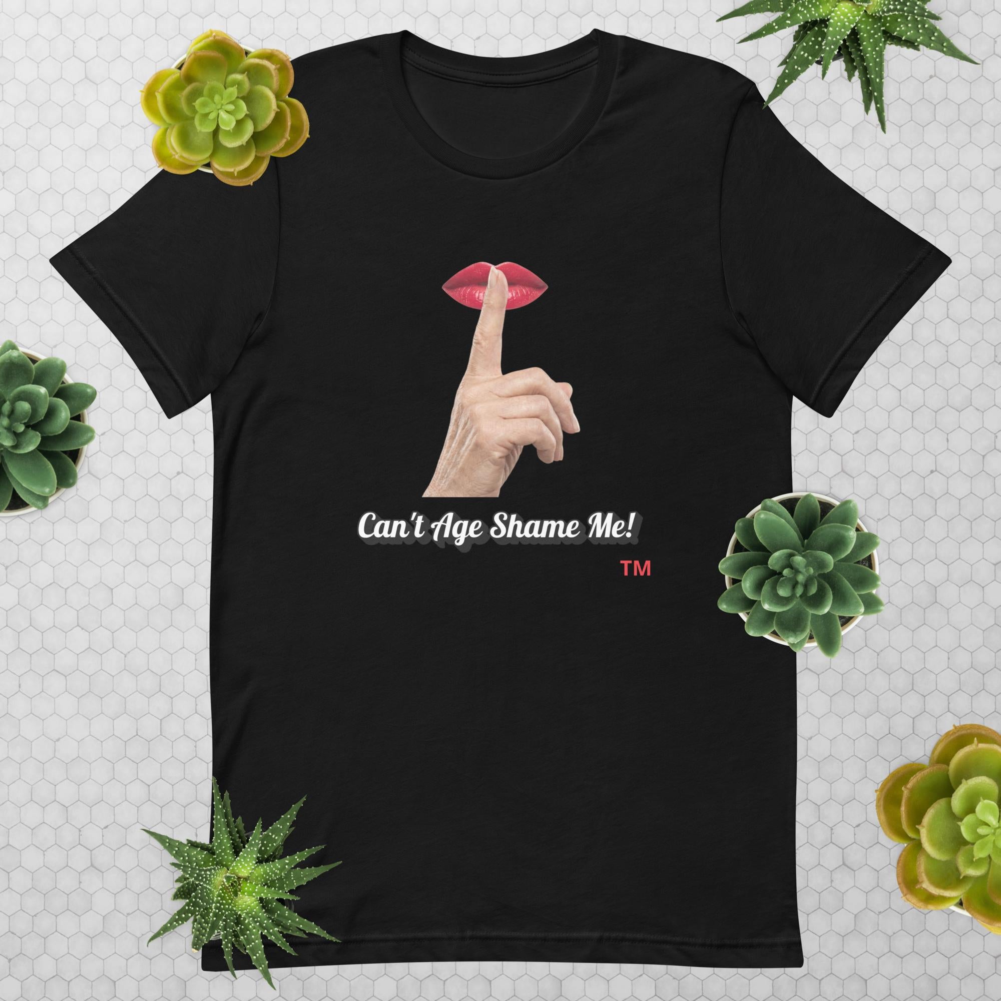 Black t-shirt with graphic design of finger on mouth and text 'CAN'T AGE SHAME ME!' text logo on a light gray background with succulents.