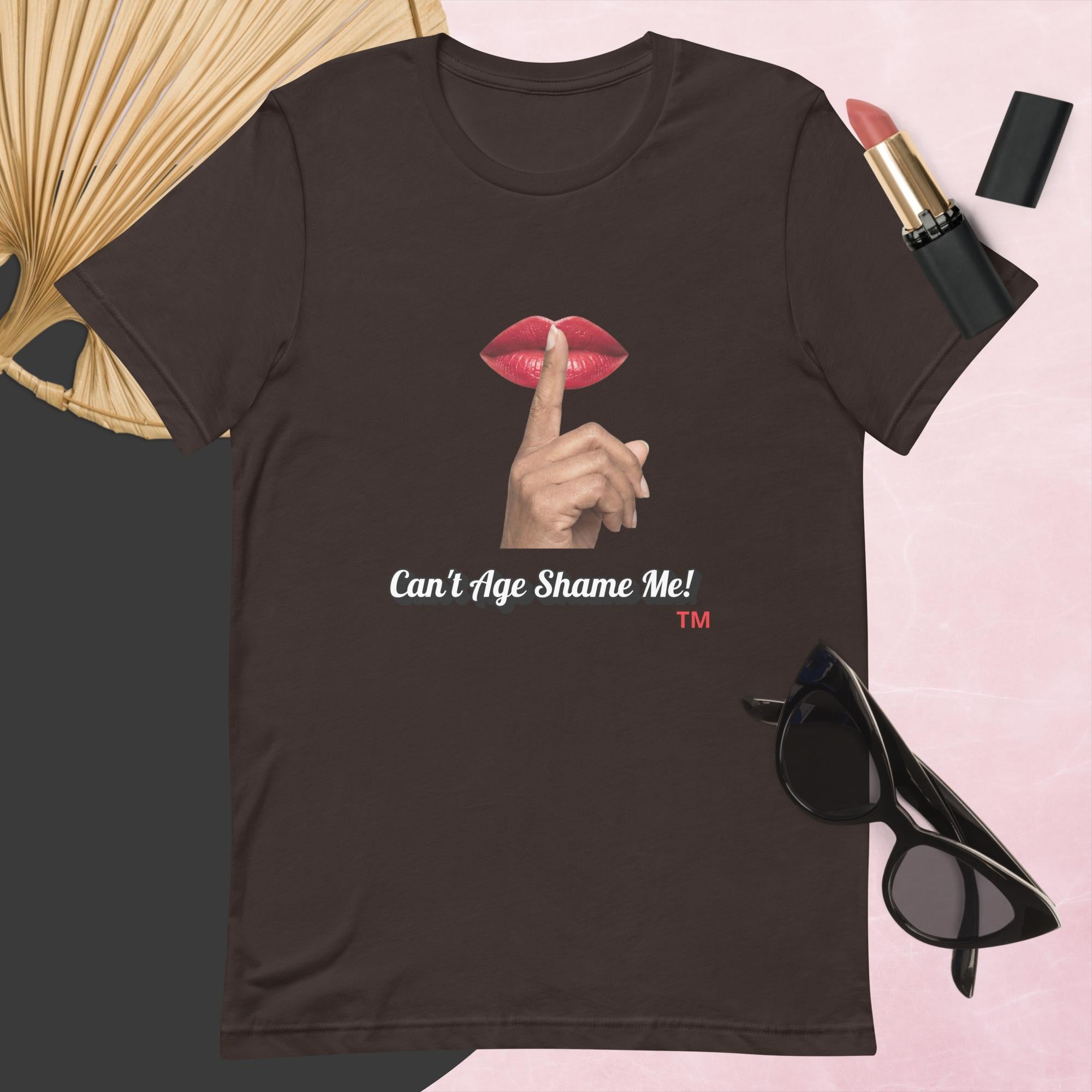 Brown t-shirt with graphic design and text, Can’t Age Shame Me!,   surrounded by sunglasses, lipstick, and a fan on a pink background.

 
