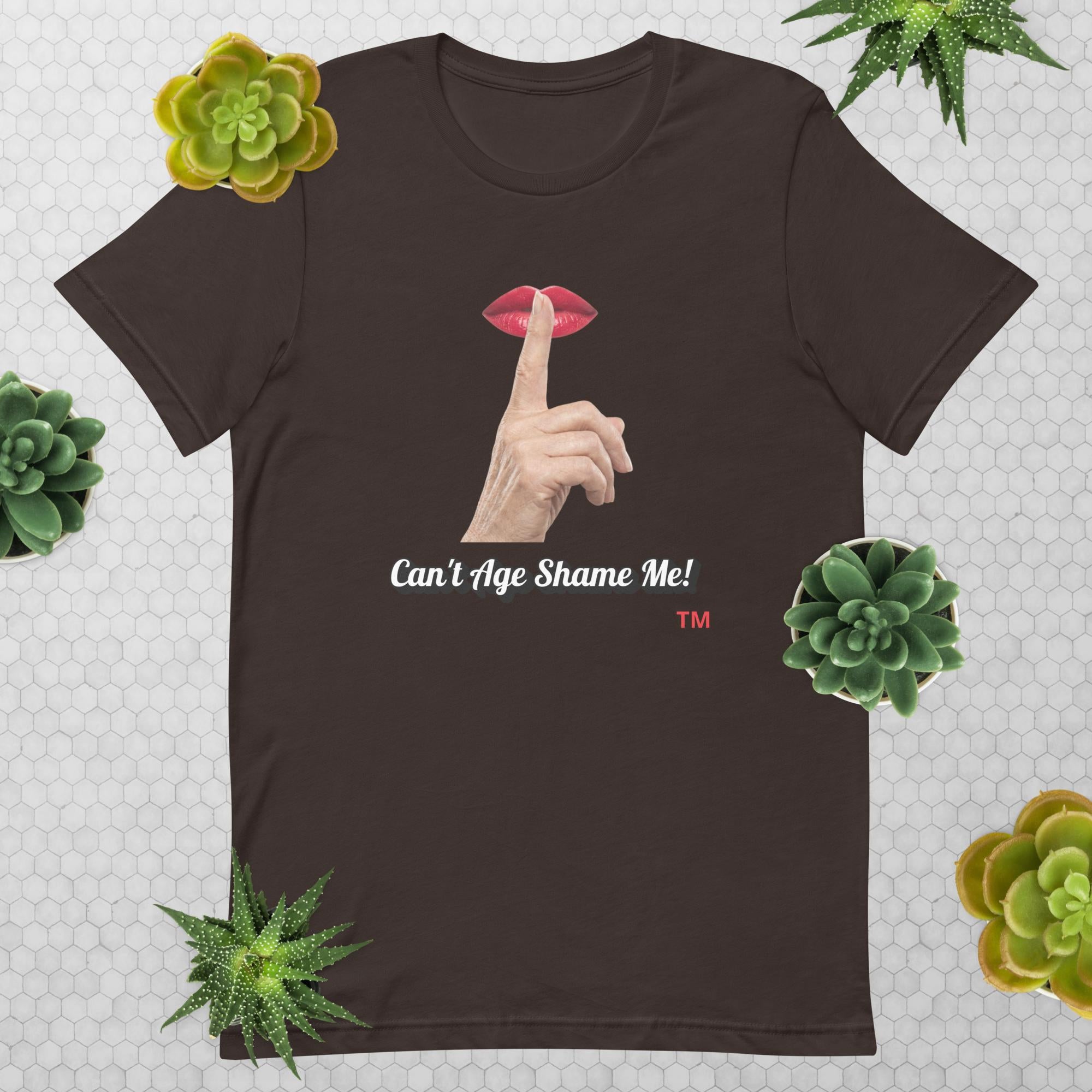 Brown t-shirt with graphic design of finger on mouth and text 'CAN'T AGE SHAME ME!' text logo on a light gray background with succulents.