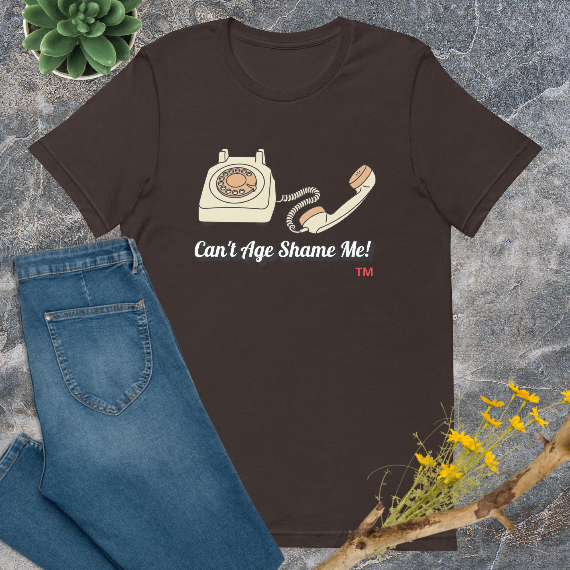 Brown t-shirt with vintage phone graphic and 'Can't Age Shame Me!' text on a stone surface with jeans and plants.