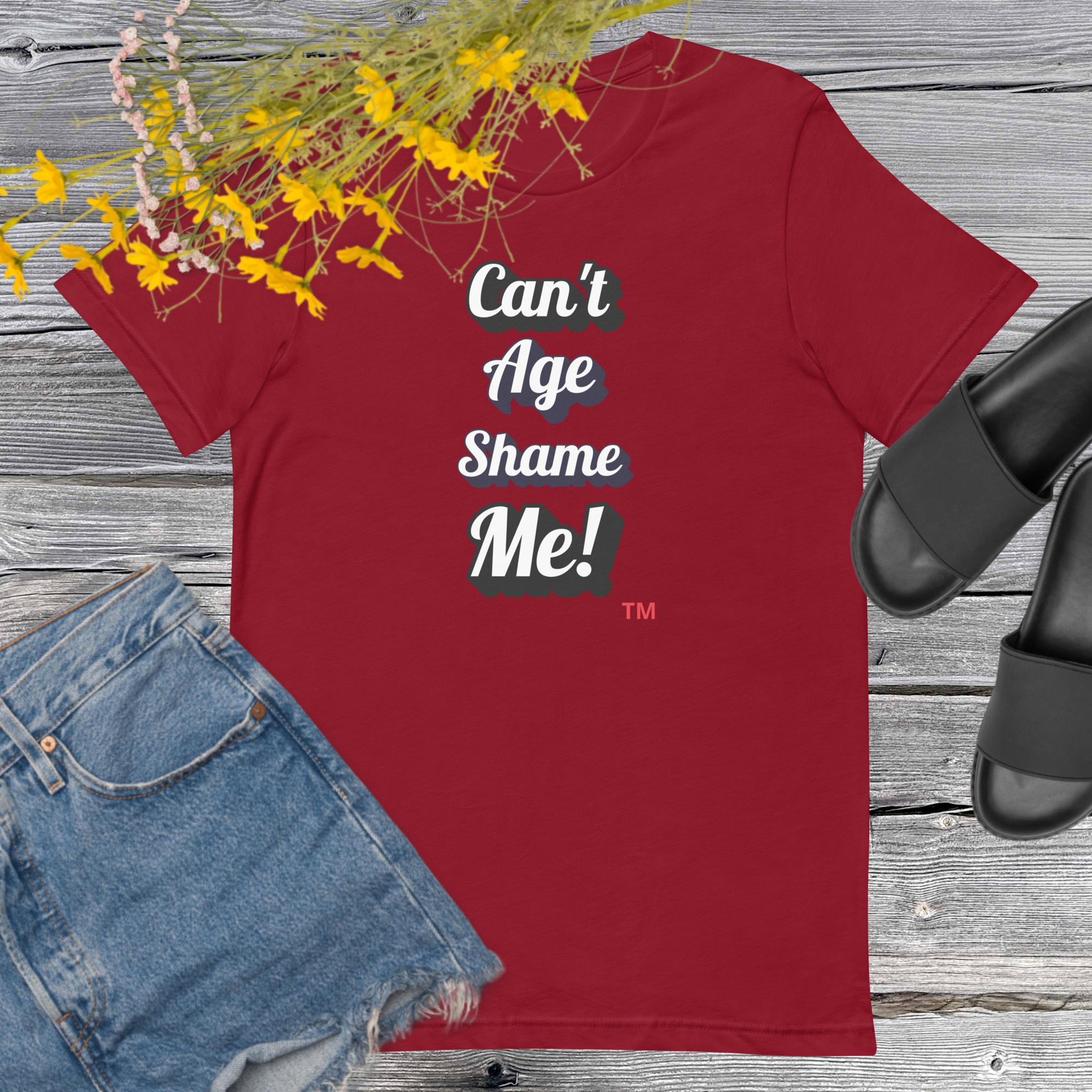 Red t-shirt with 'Can't Age Shame Me!' text on a wooden surface with denim shorts and sandals.