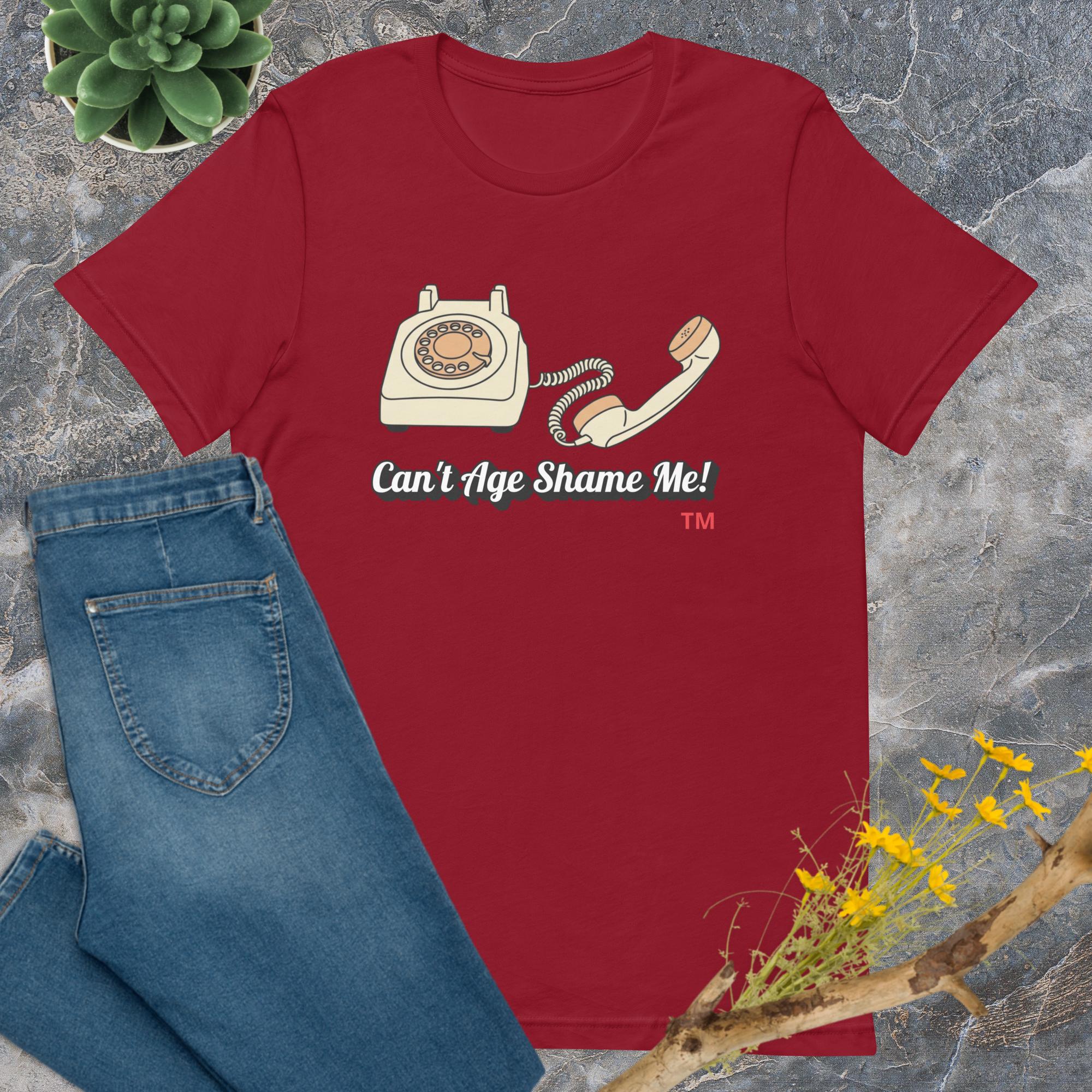 Red t-shirt with a phone graphic and text 'Can't Age Shame Me!' on a textured surface with jeans and plants.