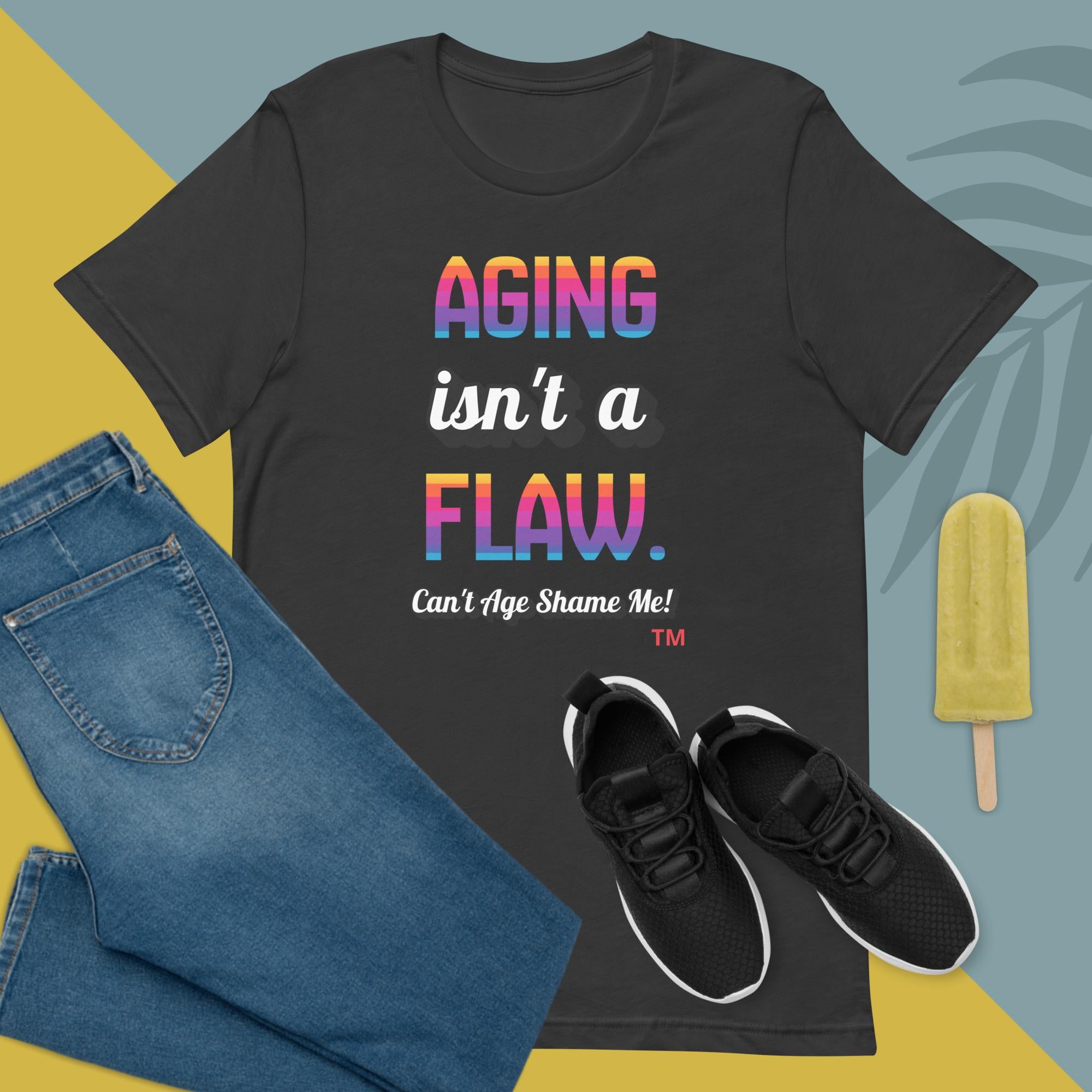 Gray t-shirt with 'Aging isn't a flaw' text, paired with jeans and sneakers on a colorful background.
