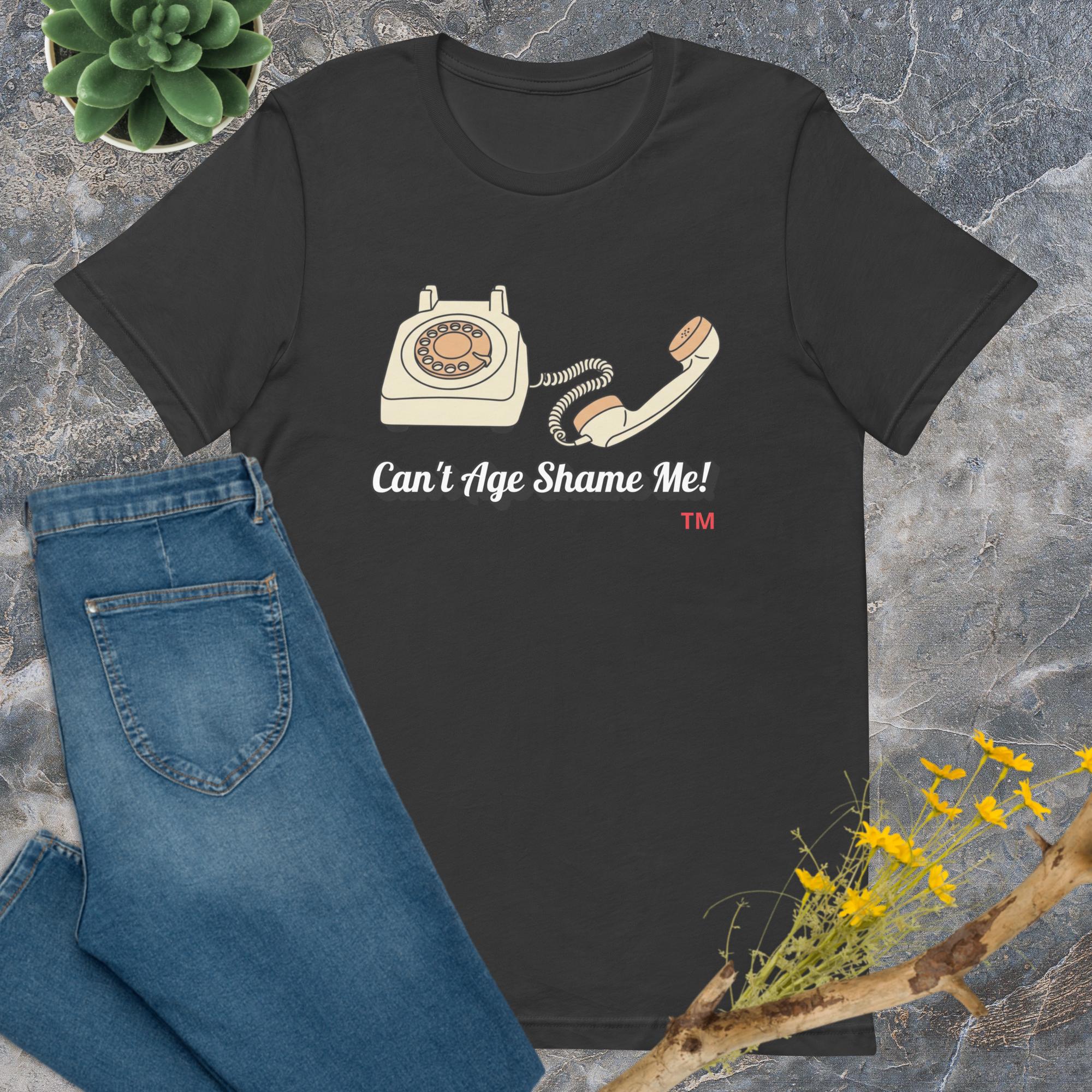 dark gray t-shirt with phone graphic and text 'Can't Age Shame Me!' on a stone surface with jeans and flowers.