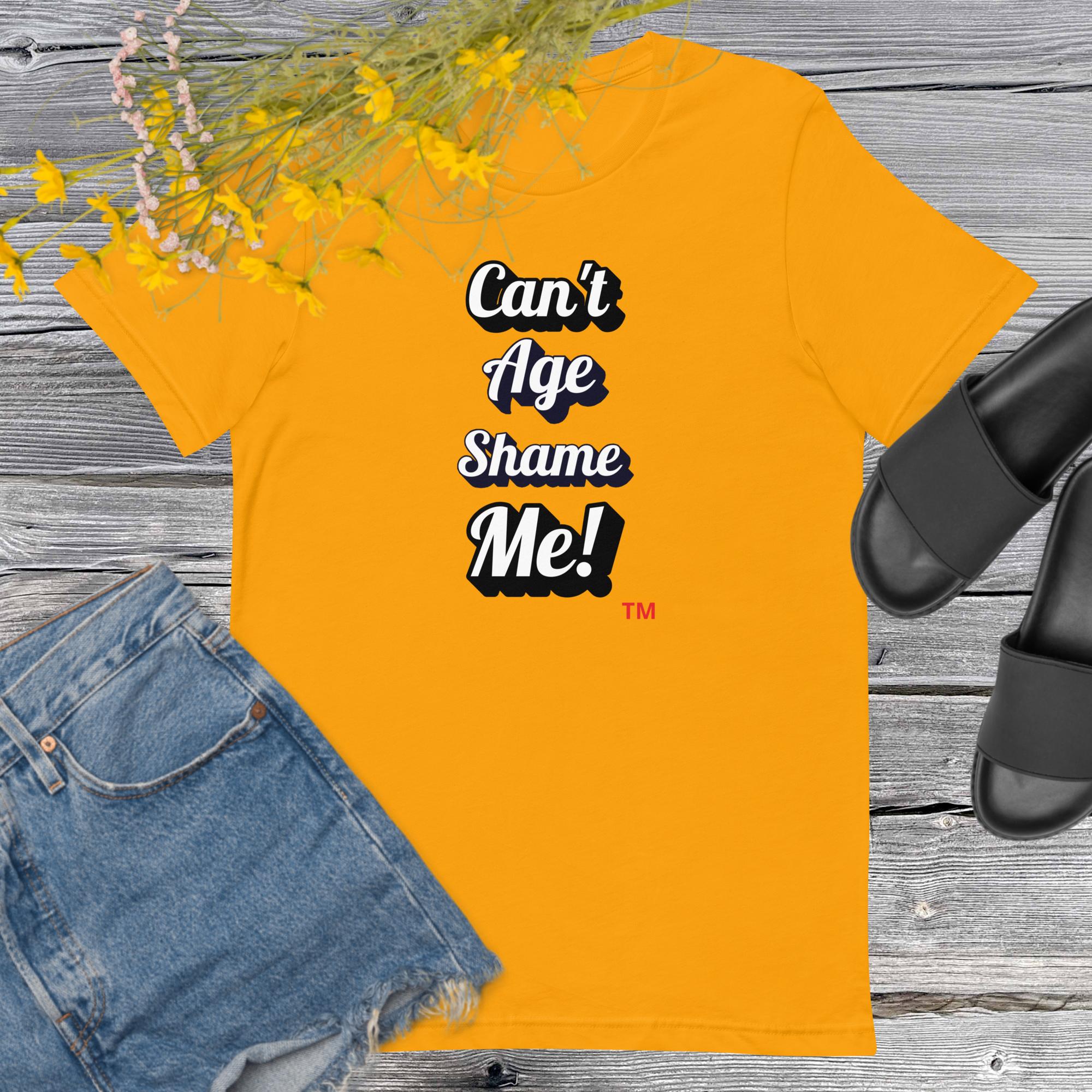 Yellow t-shirt with 'Can't Age Shame Me!' text on a wooden surface with denim shorts and sandals.