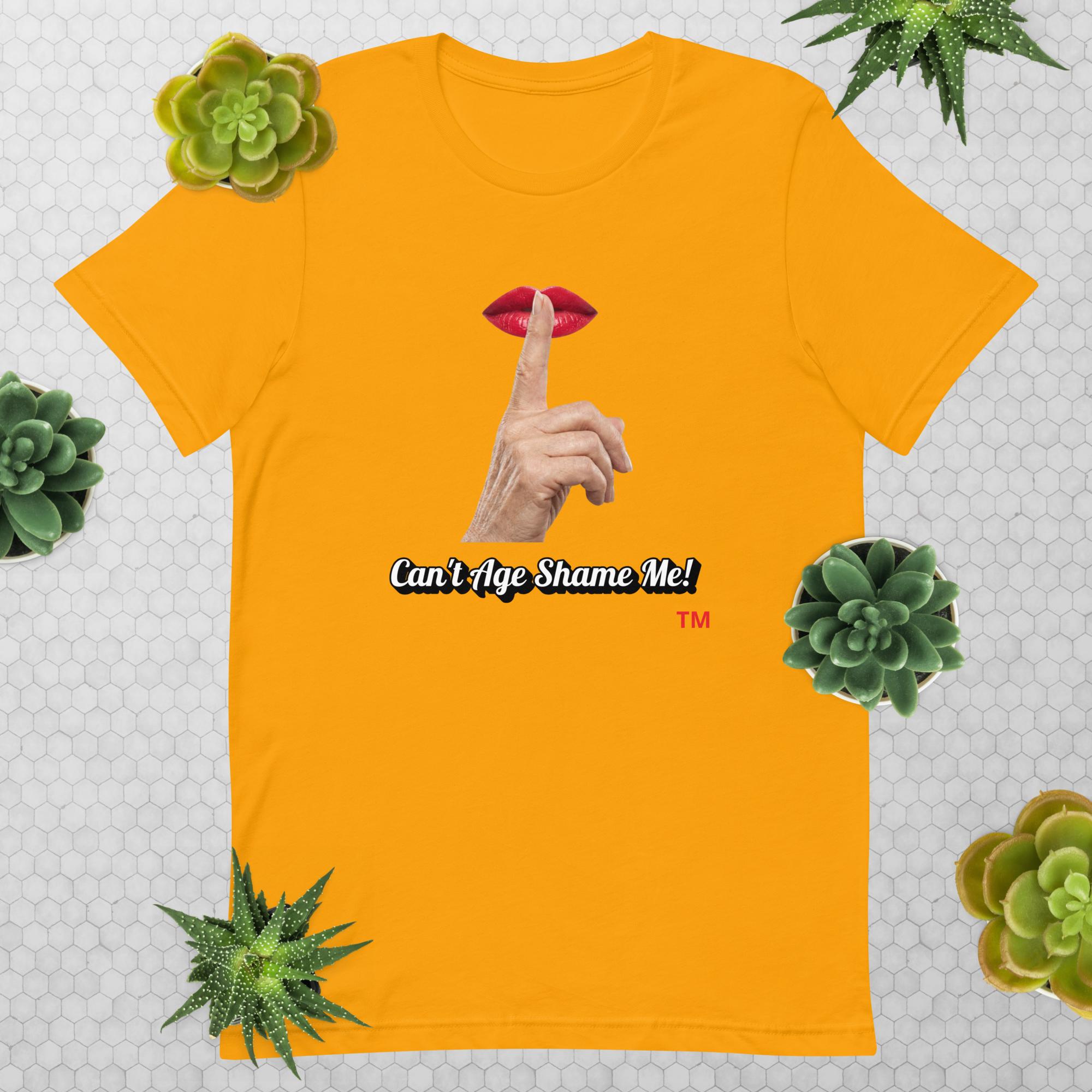 Gold t-shirt with graphic design of finger on mouth and text 'CAN'T AGE SHAME ME!' text logo on a light gray background with succulents.
