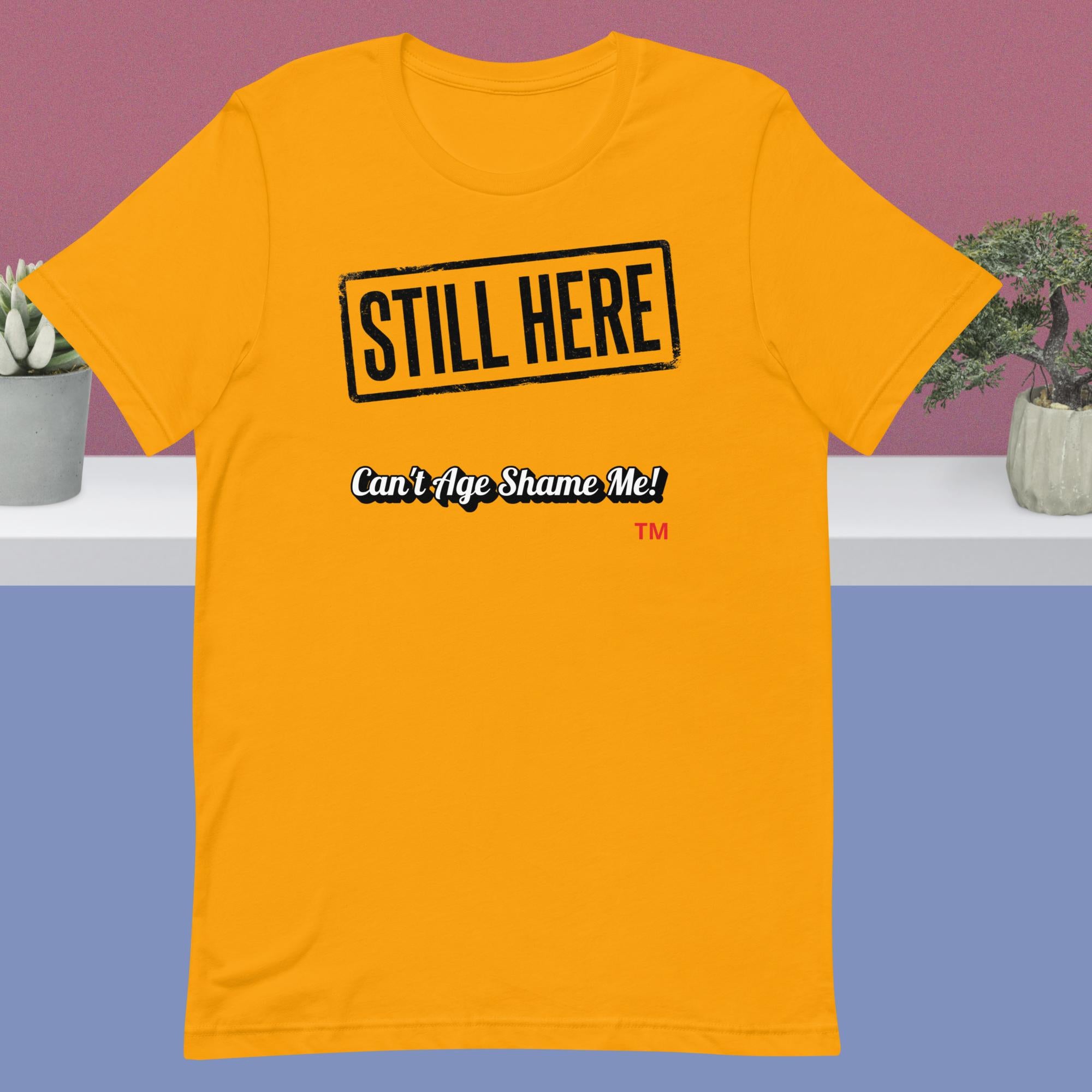 Gold  t-shirt with 'STILL HERE Can't Age Shame Me' text on a purple and blue gradient background.