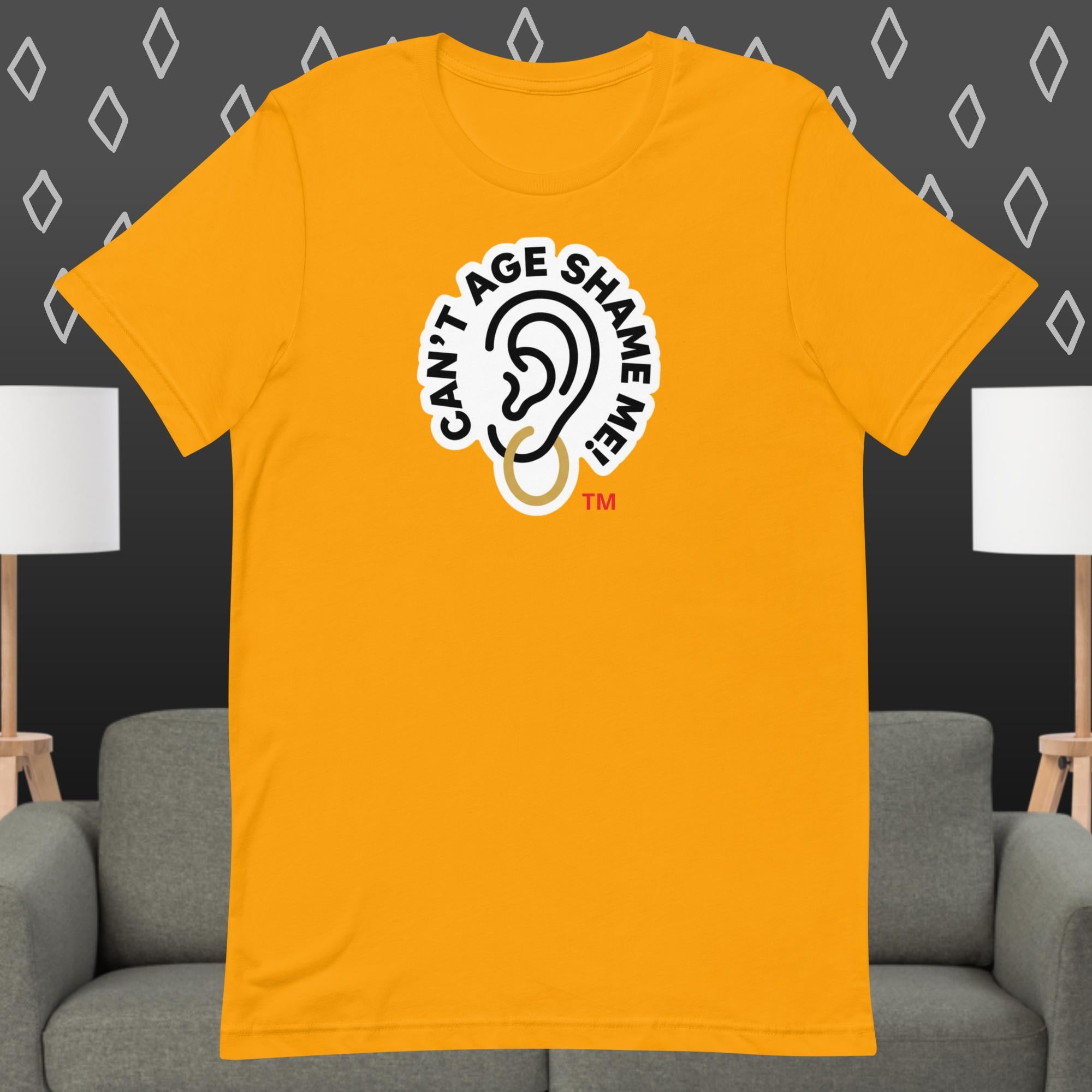 Gold t-shirt with 'Can't Age Shame Me' graphic on a gray couch and lamp background