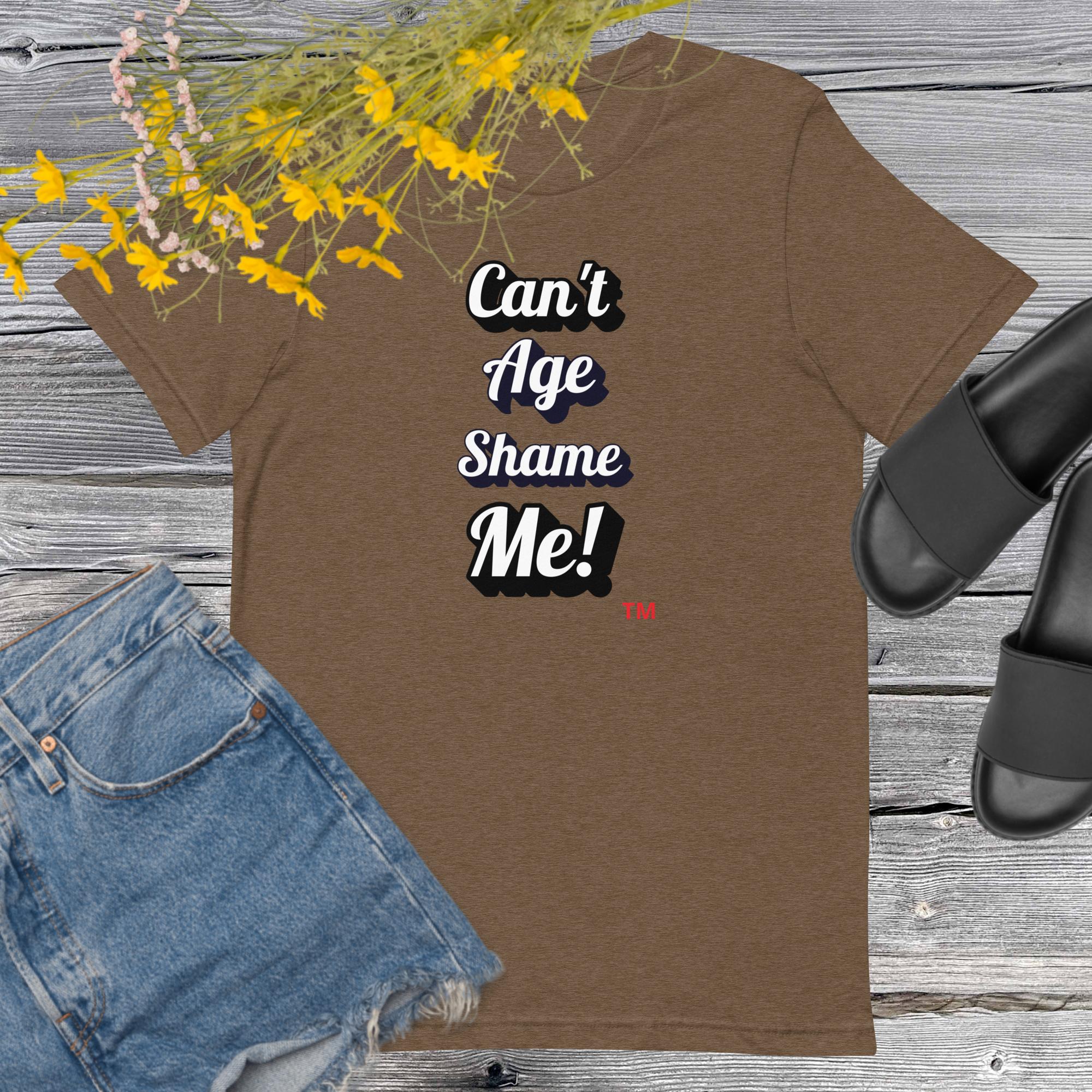 Brown t-shirt with 'Can't Age Shame Me!' text, paired with denim shorts and black sandals on a wooden surface.