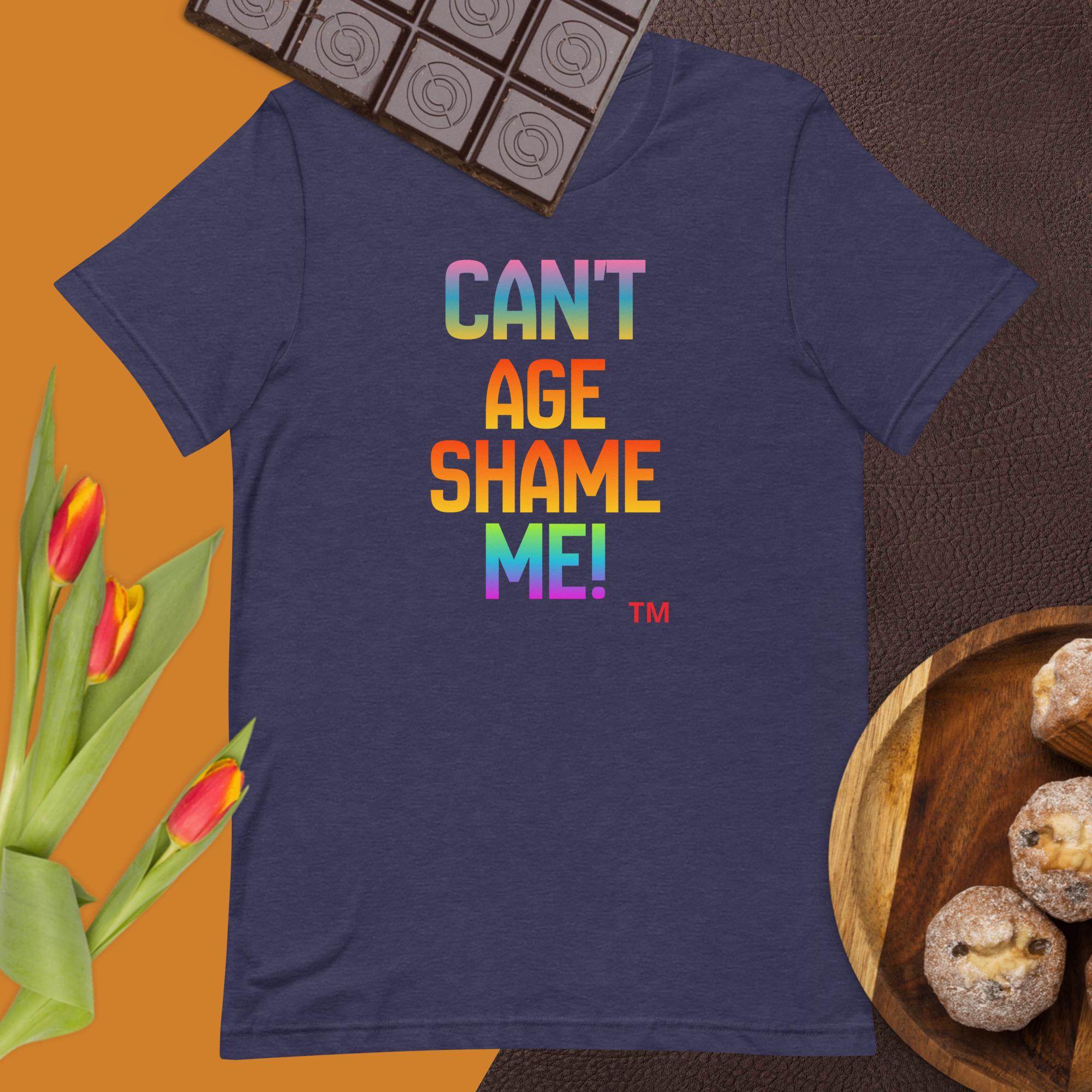 Navy blue t-shirt with colorful text 'CAN'T AGE SHAME ME!' on a brown surface with flowers and cookies.