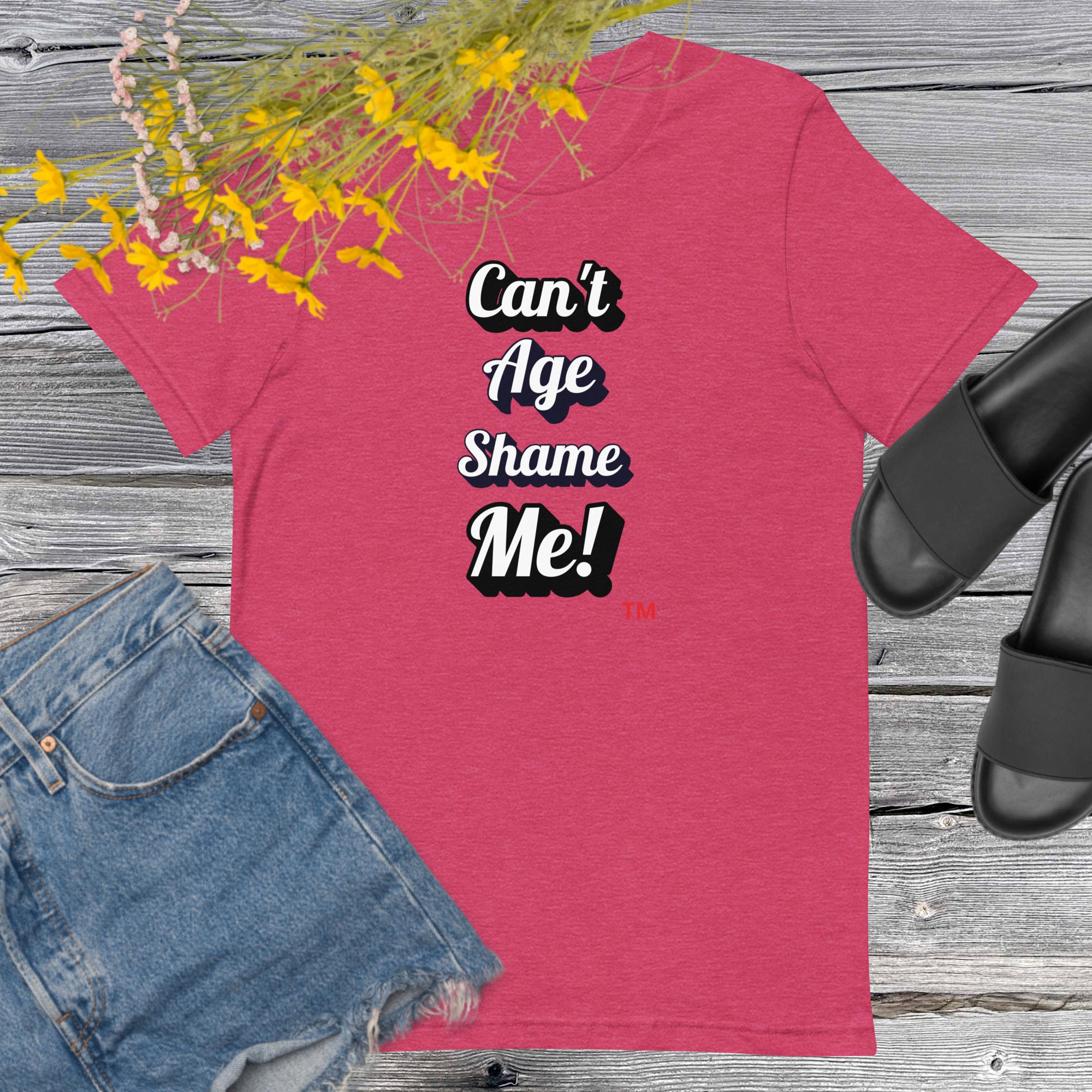 Pink t-shirt with 'Can't Age Shame Me!' text, paired with denim shorts and black sandals on a wooden surface.