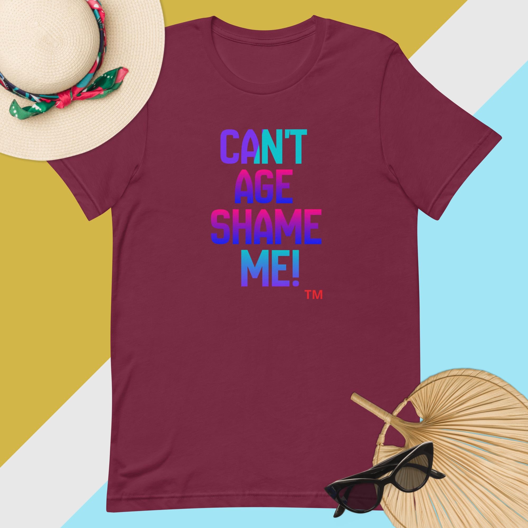 Maroon t-shirt with colorful text 'CAN'T AGE SHAME ME!' on a multicolored background with accessories.