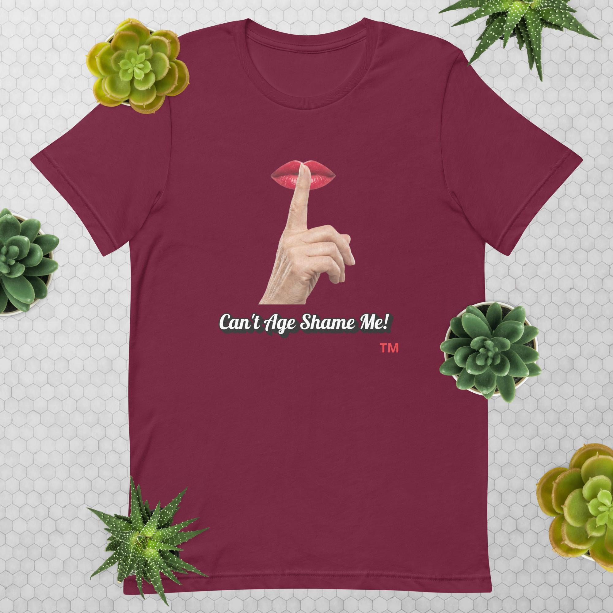 Maroon t-shirt with graphic design of finger on mouth and text 'CAN'T AGE SHAME ME!' text logo on a light gray background with succulents.