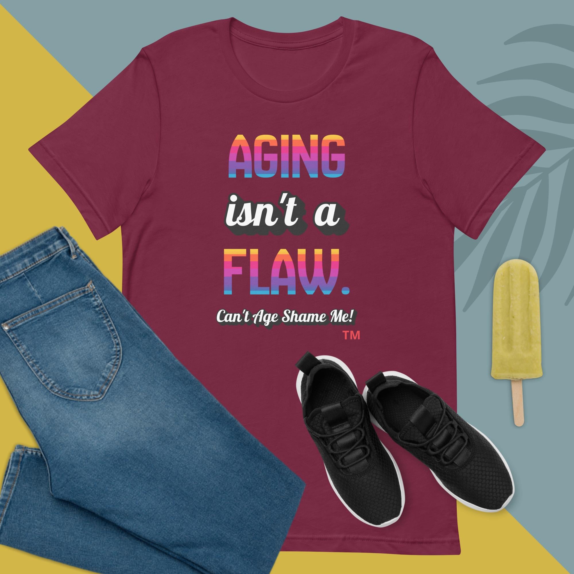 Maroon  t-shirt with 'Aging isn't a flaw' text, paired with jeans and sneakers on a colorful background.
