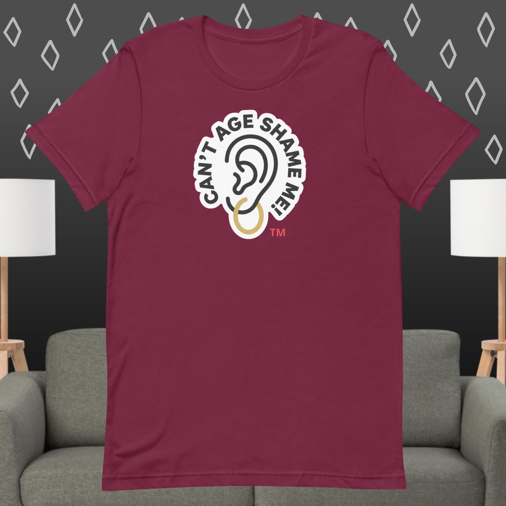 Maroon t-shirt with 'Can't Age Shame Me' graphic on a gray couch and lamp background