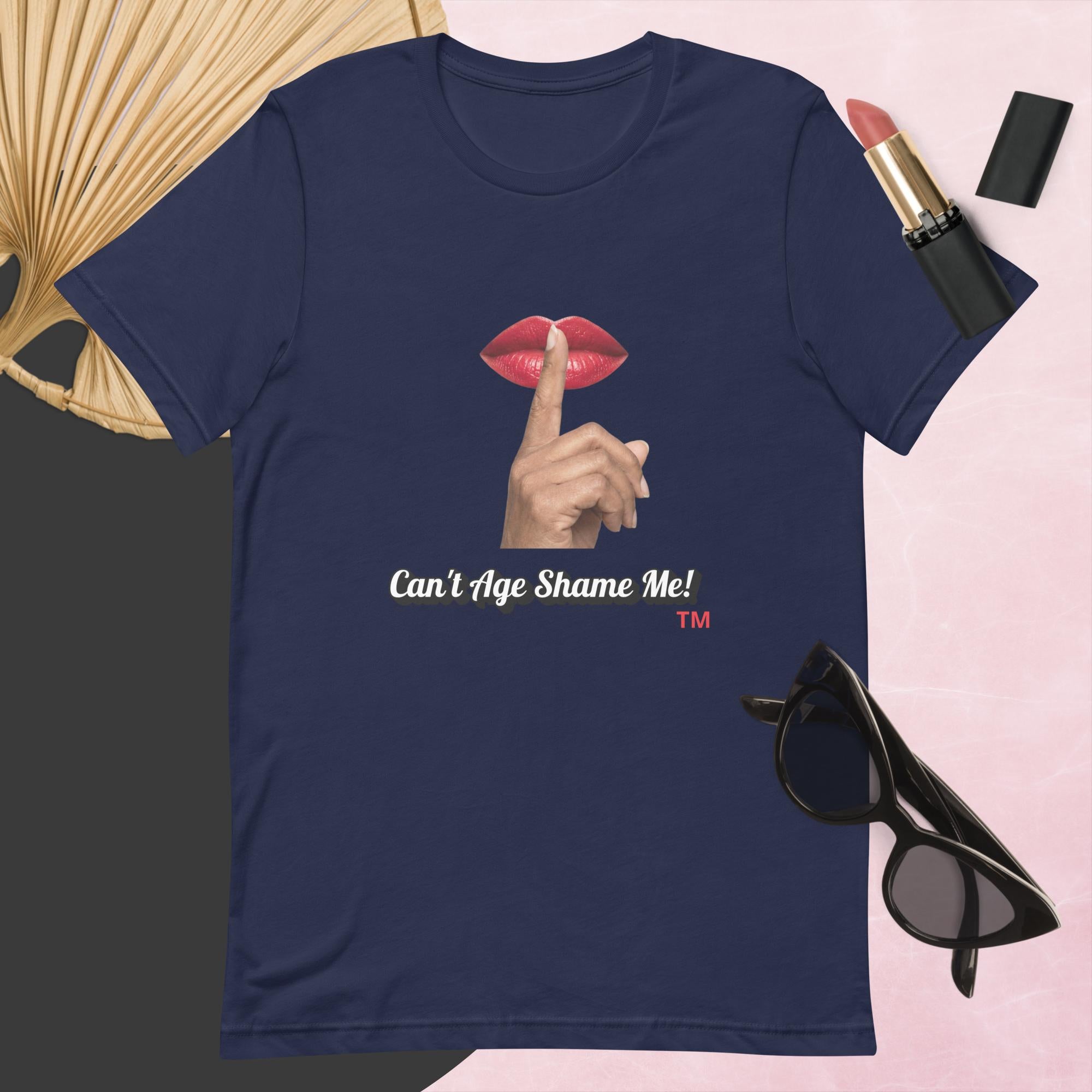 Navy t-shirt with graphic design and text, Can’t Age Shame Me!,   surrounded by sunglasses, lipstick, and a fan on a pink background.

 
