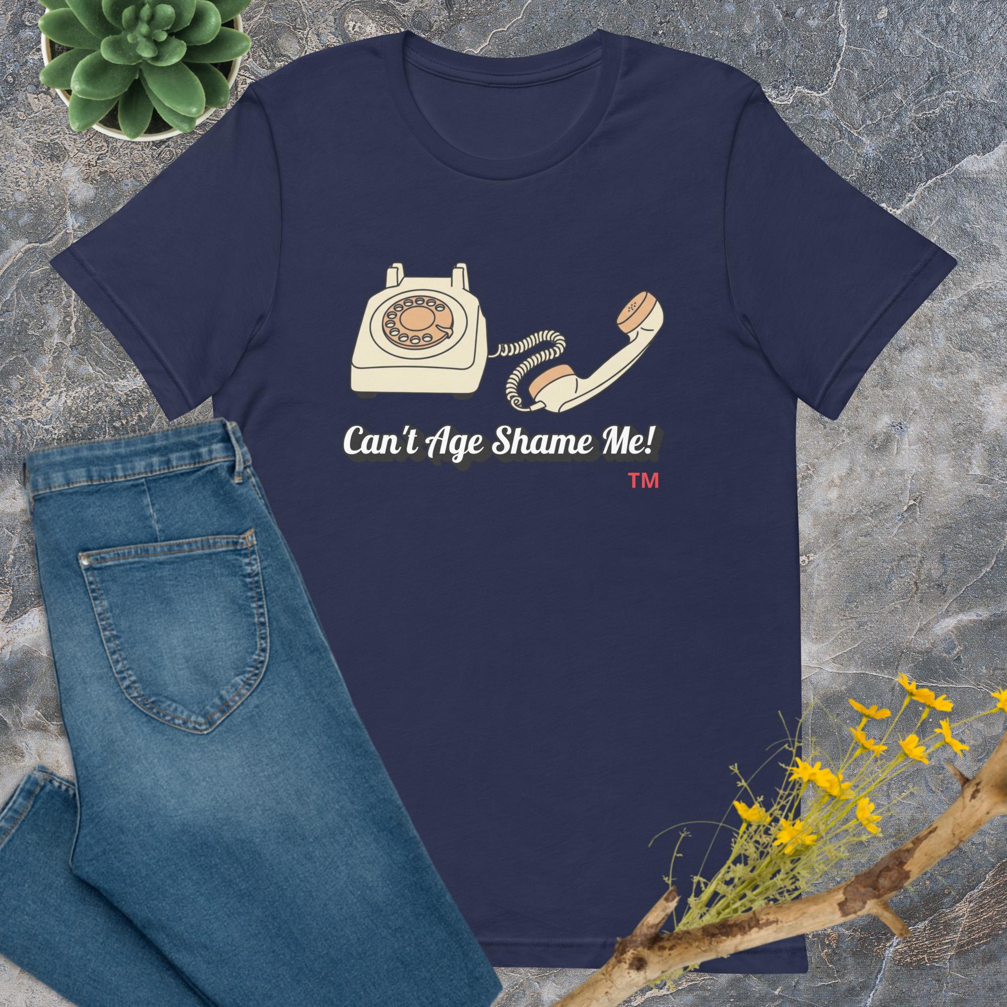 Navy blue t-shirt with vintage phone graphic and 'Can't Age Shame Me!' text on a textured surface with jeans and flowers.