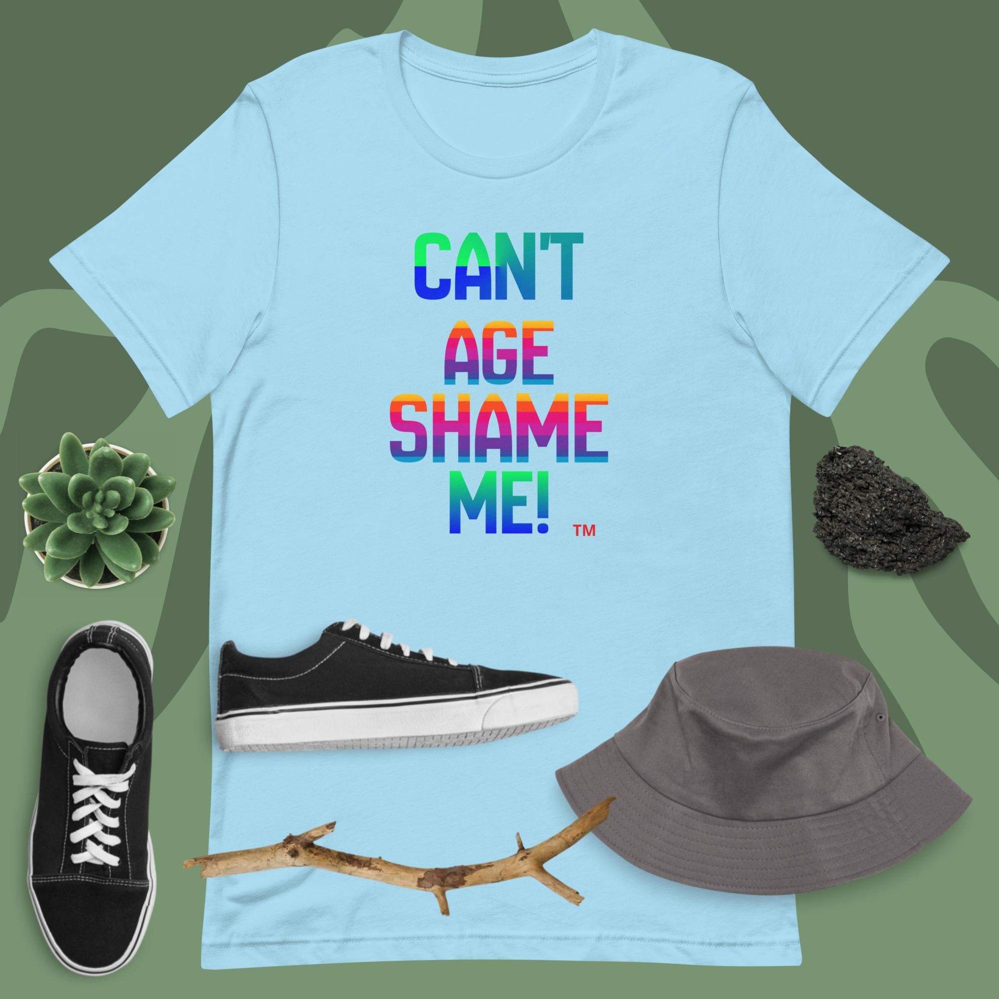 Light blue t-shirt with colorful text 'CAN'T AGE SHAME ME!' on a green background with shoes, a hat, and a branch.