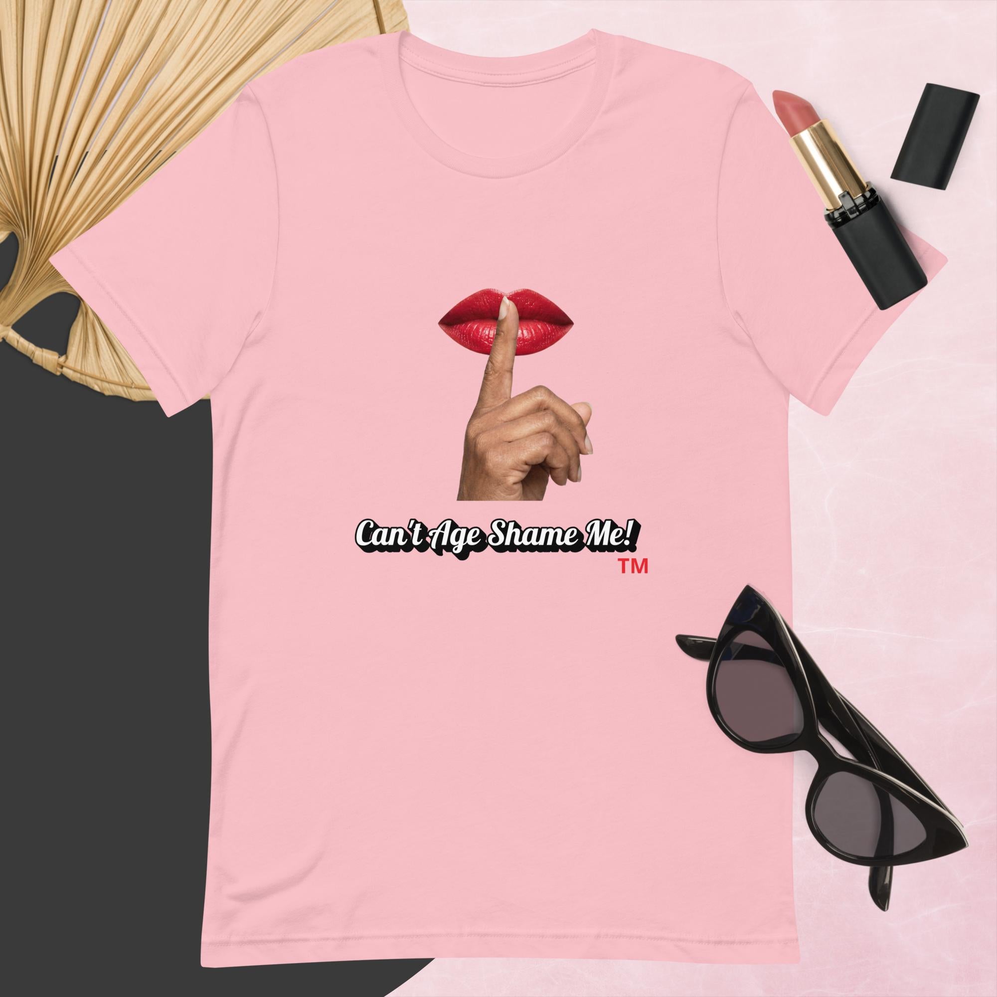 Pink t-shirt with graphic design and text, Can’t Age Shame Me!,   surrounded by sunglasses, lipstick, and a fan on a pink background.

 
