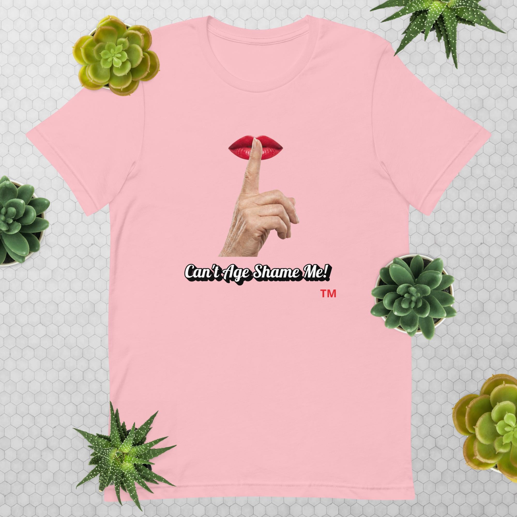 Pink t-shirt with graphic design of finger on mouth and text 'CAN'T AGE SHAME ME!' text logo on a light gray background with succulents.