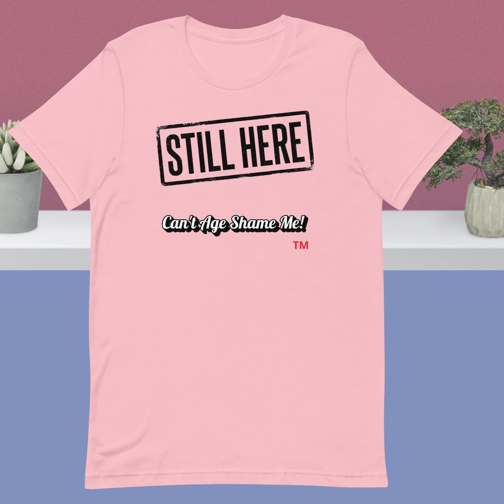 Pink t-shirt with 'STILL HERE Can't Age Shame Me' text on a purple background