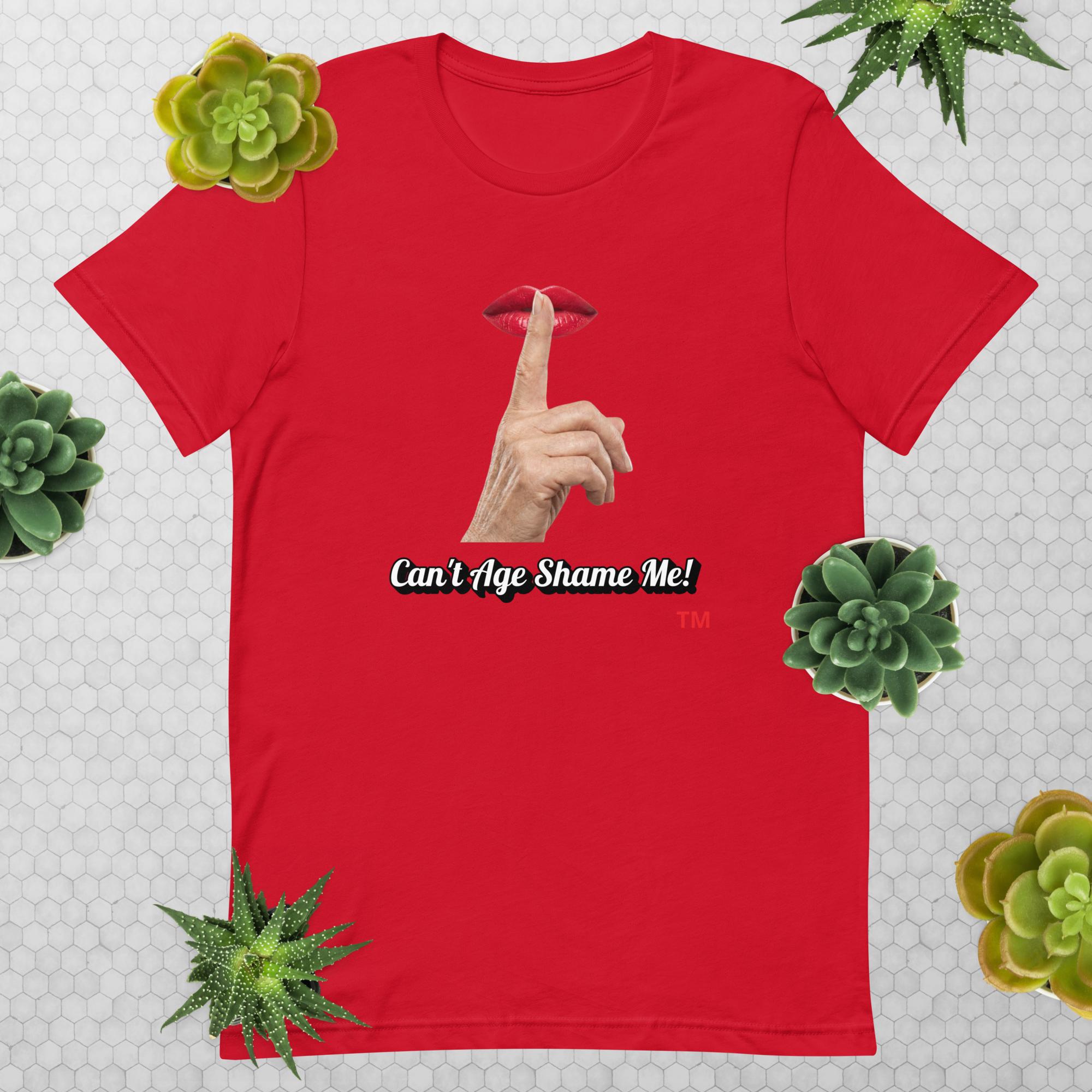 Red t-shirt with graphic design of finger on mouth and text 'CAN'T AGE SHAME ME!' text logo on a light gray background with succulents.