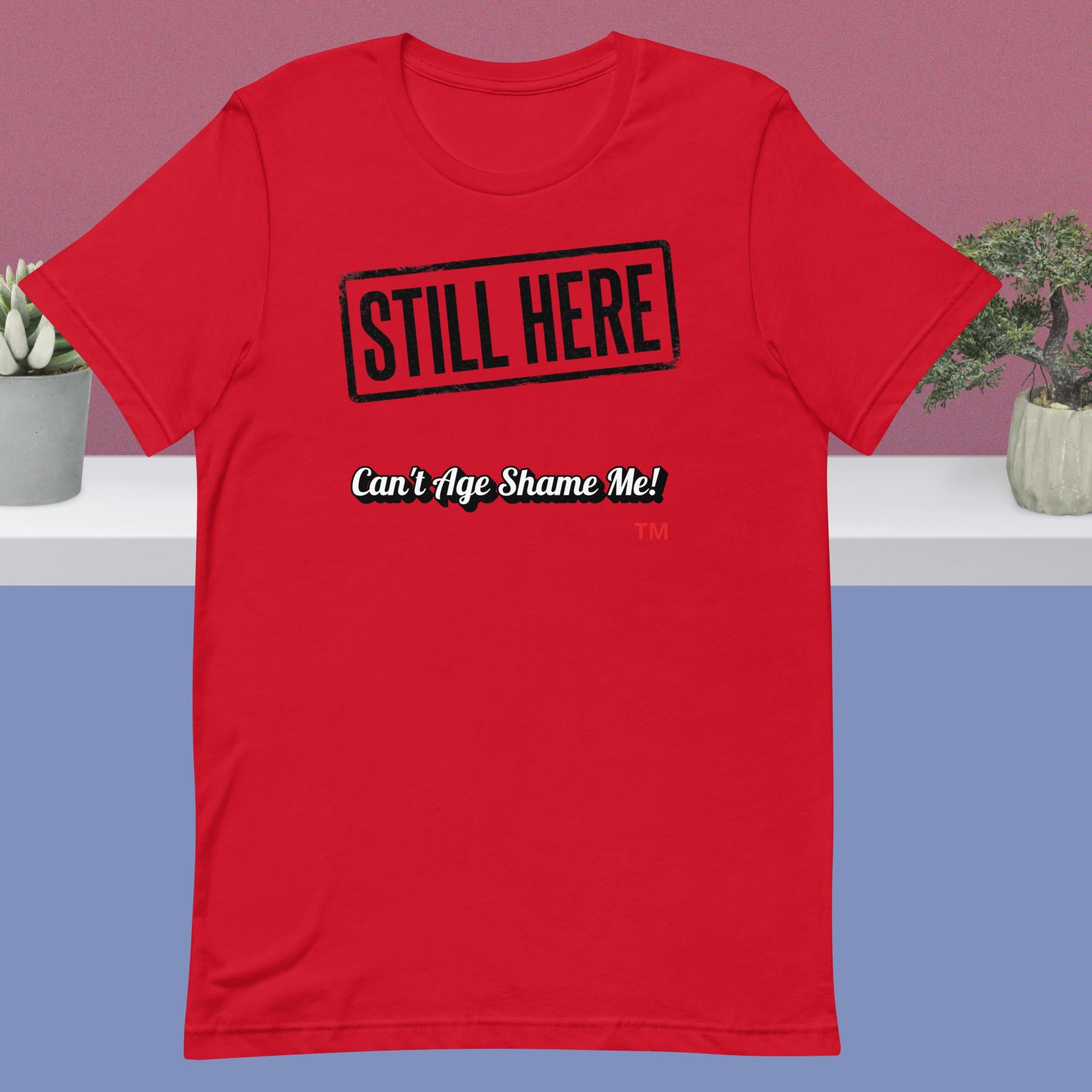 Red t-shirt with 'STILL HERE Can't Age Shame Me!' text on a plain background