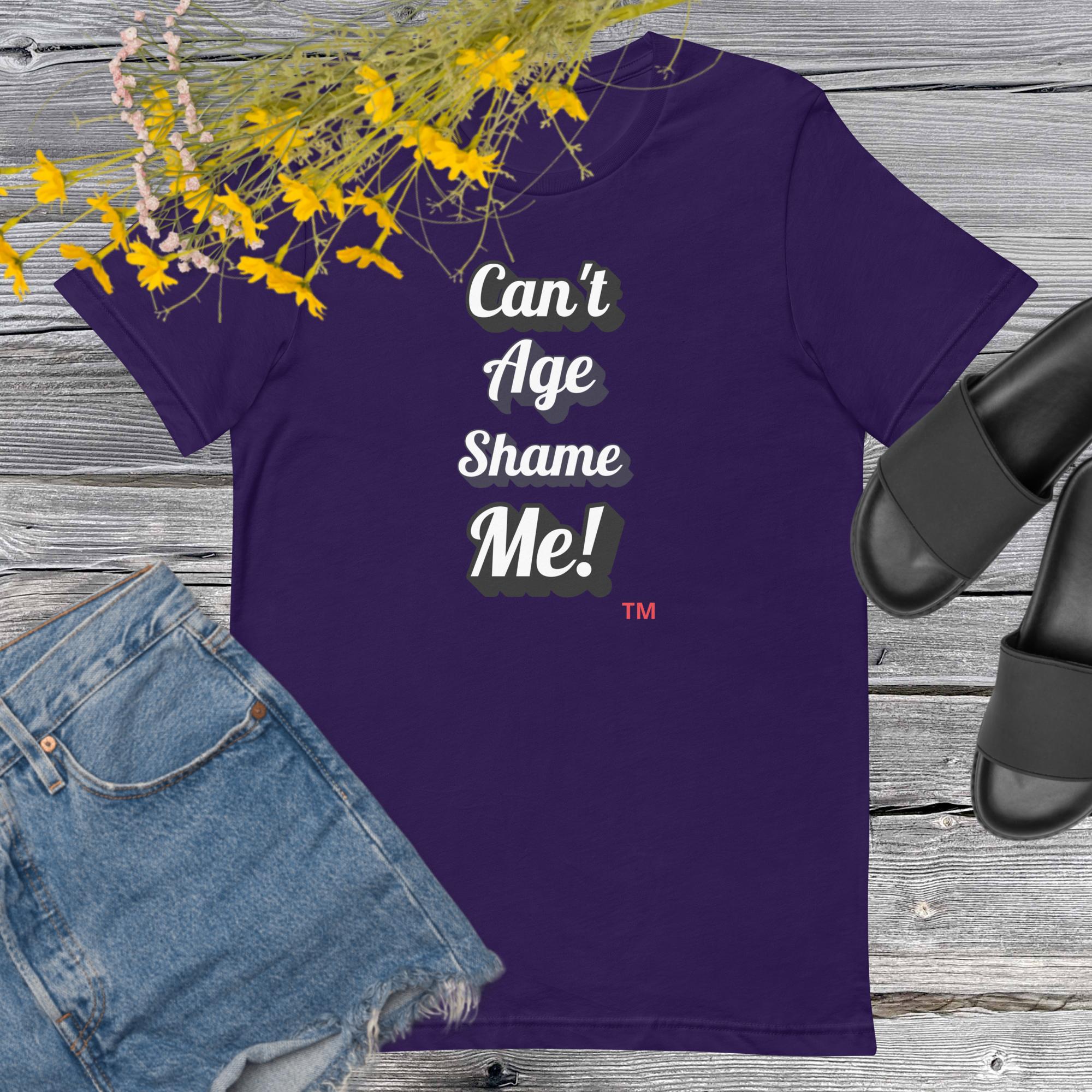 Purple t-shirt with 'Can't Age Shame Me!' text on a wooden surface with denim shorts and black sandals.