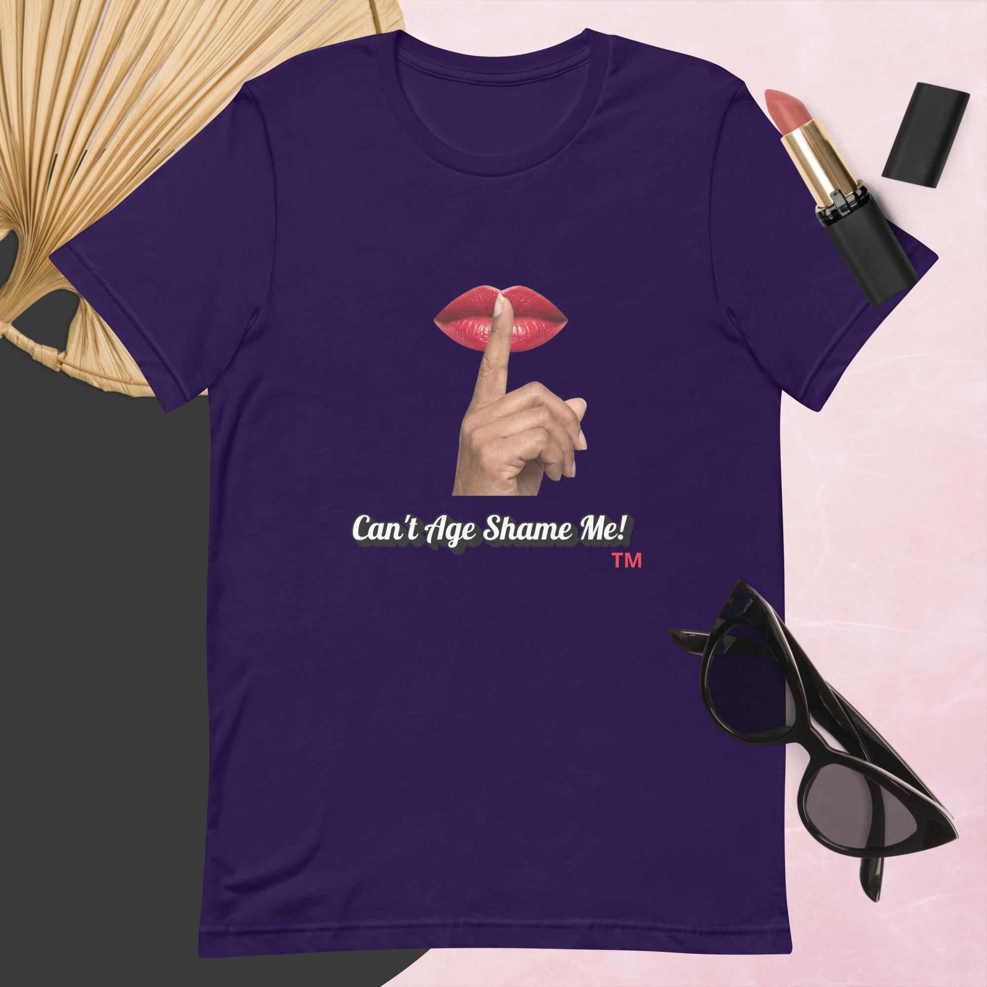 Purple t-shirt with graphic design and text, Can’t Age Shame Me!,   surrounded by sunglasses, lipstick, and a fan on a pink background.

 

