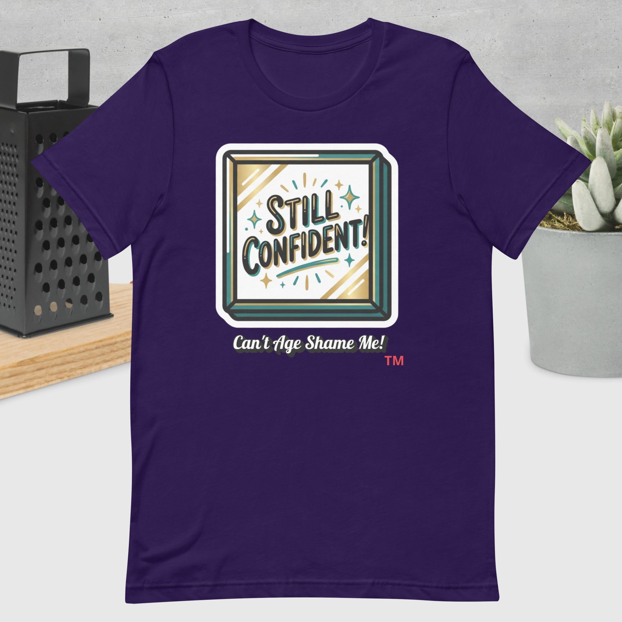 Purple t-shirt with 'Still Confident! Can't Age Shame Me!' graphic design on a neutral background.