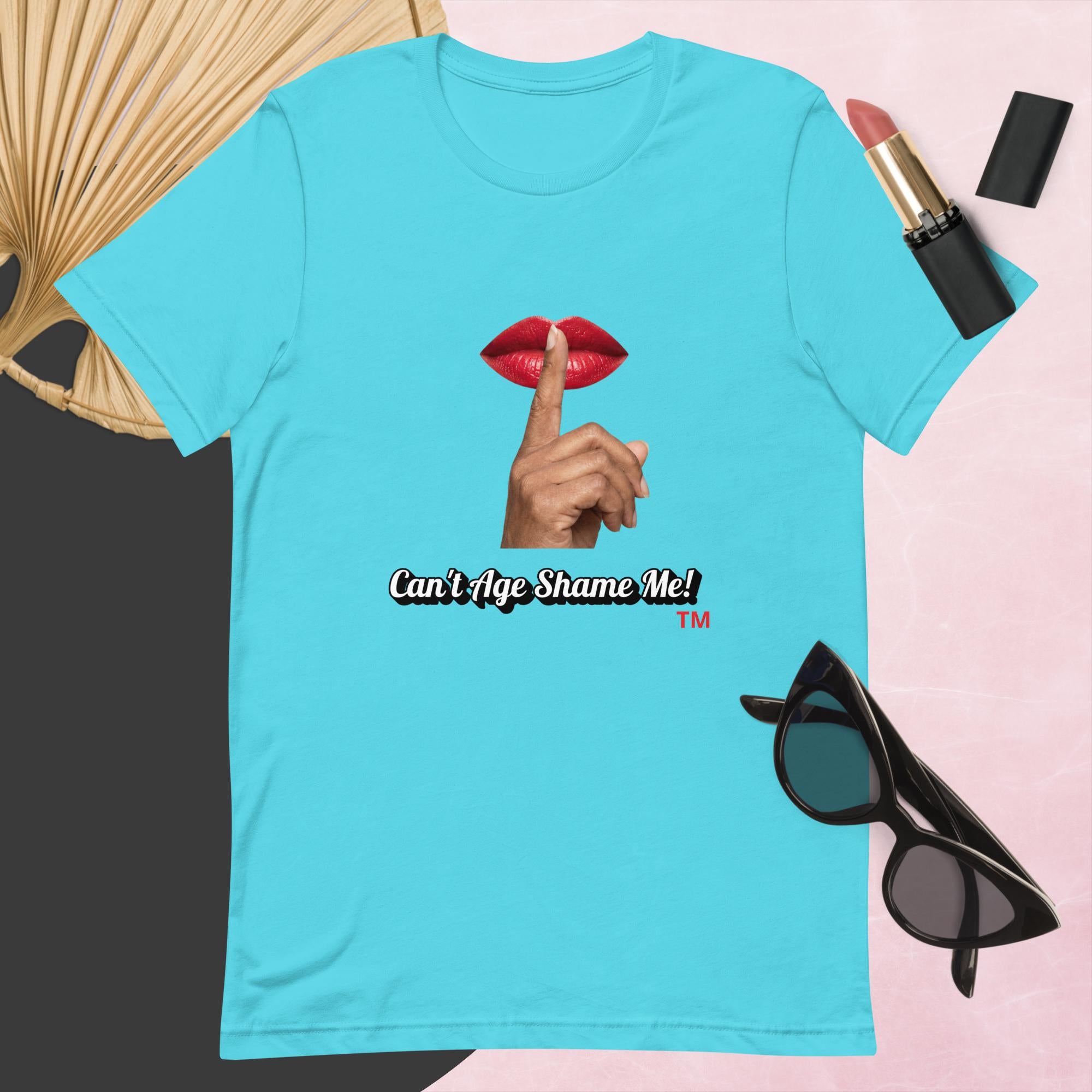 Turquoise t-shirt with graphic design and text, Can’t Age Shame Me!,   surrounded by sunglasses, lipstick, and a fan on a pink background.

 

