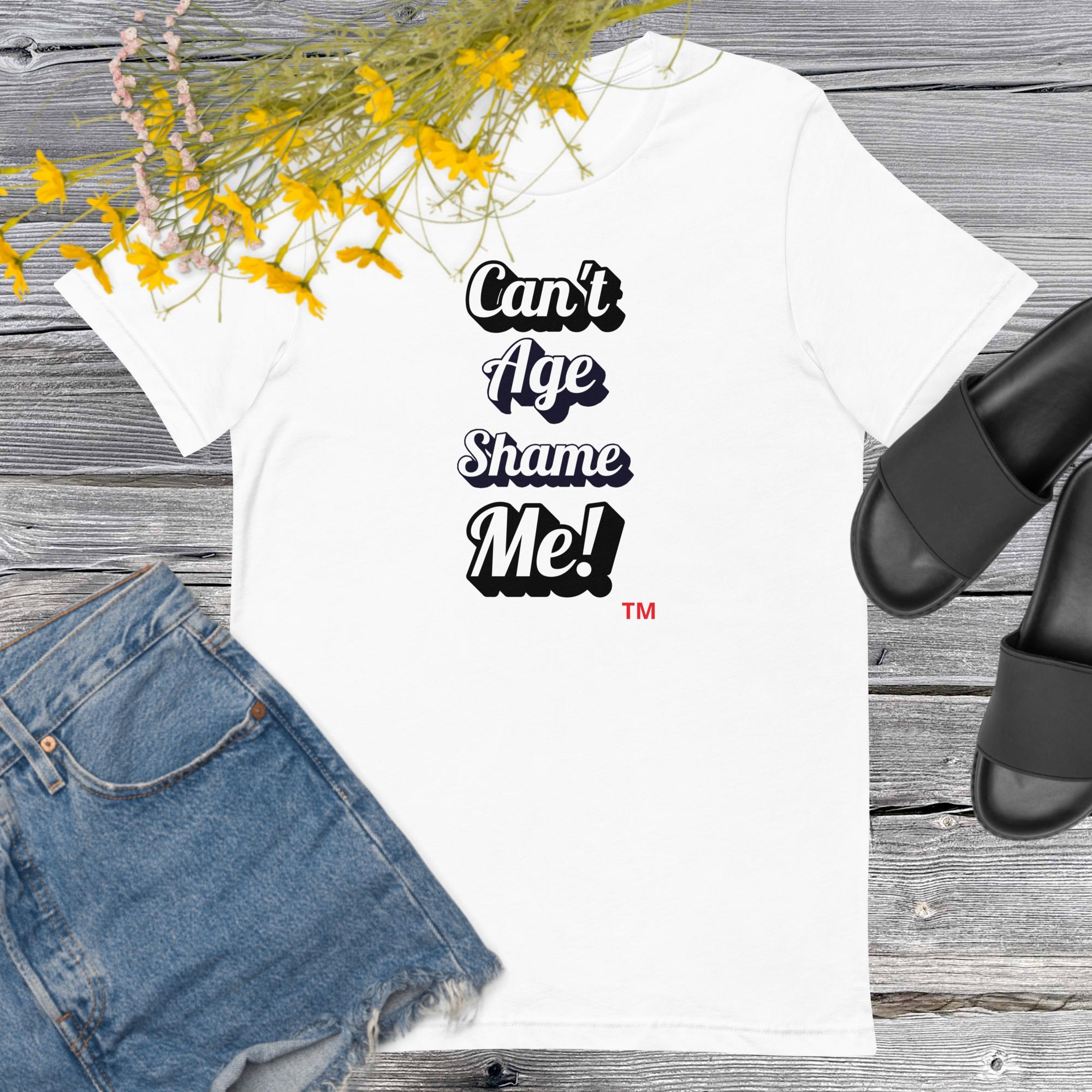 White t-shirt with 'Can't Age Shame Me!' text, denim shorts, and black sandals on a wooden surface with yellow flowers.
