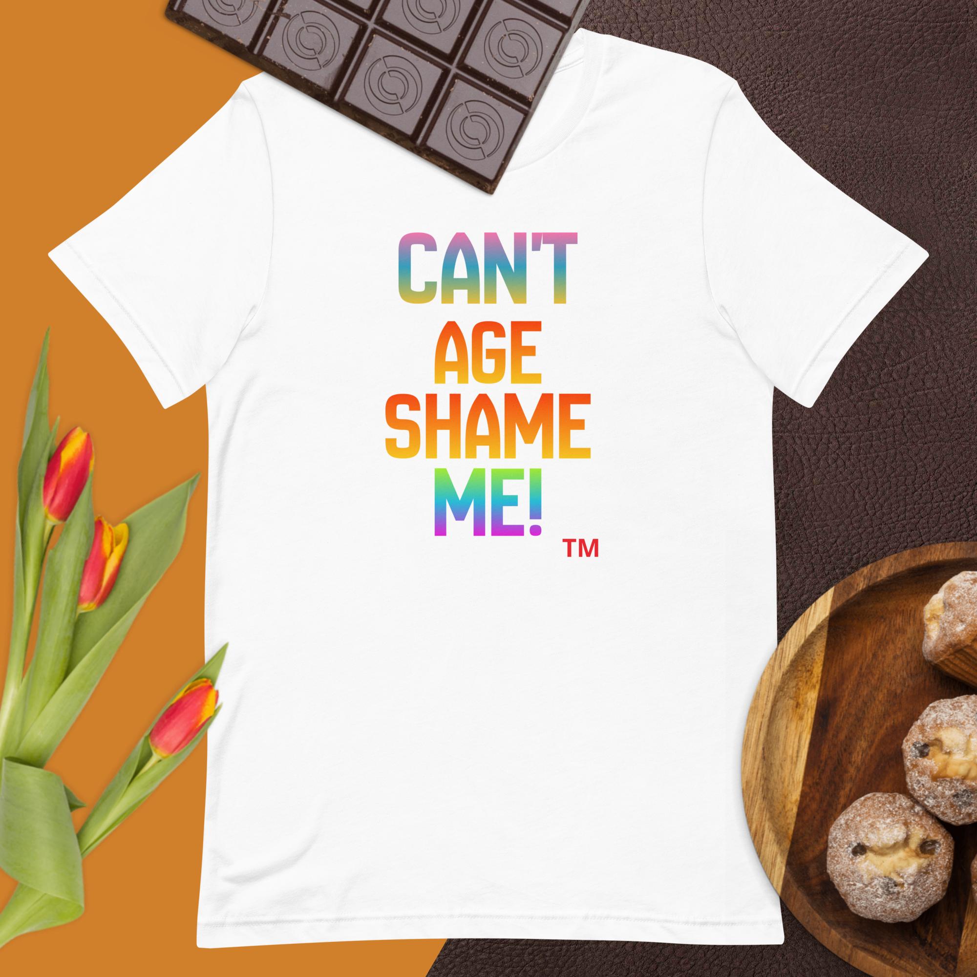 White t-shirt with colorful text 'CAN'T AGE SHAME ME!' on a brown surface with flowers and chocolate.
