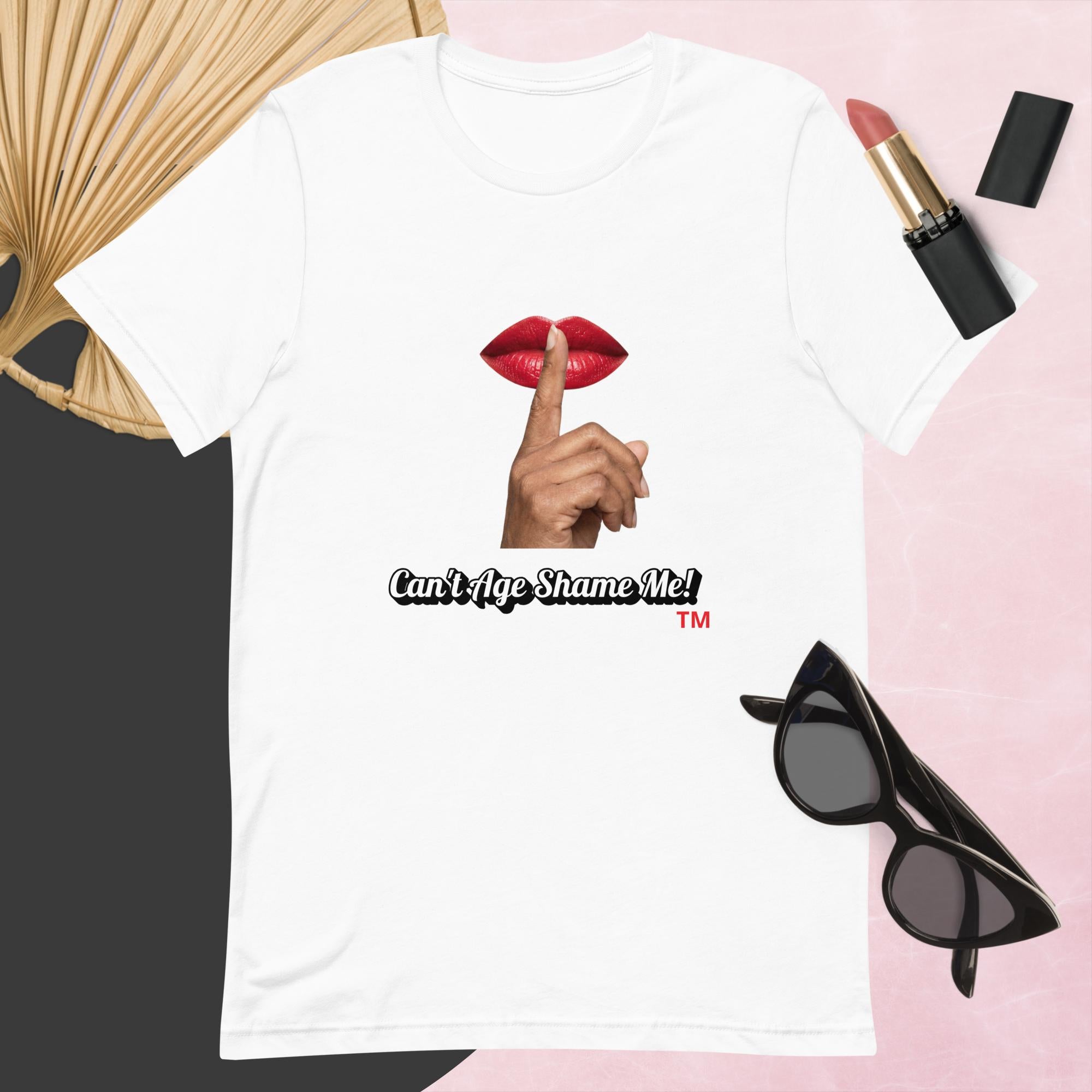 White t-shirt with graphic design and text, Can’t Age Shame Me!,   surrounded by sunglasses, lipstick, and a fan on a pink background.

 
